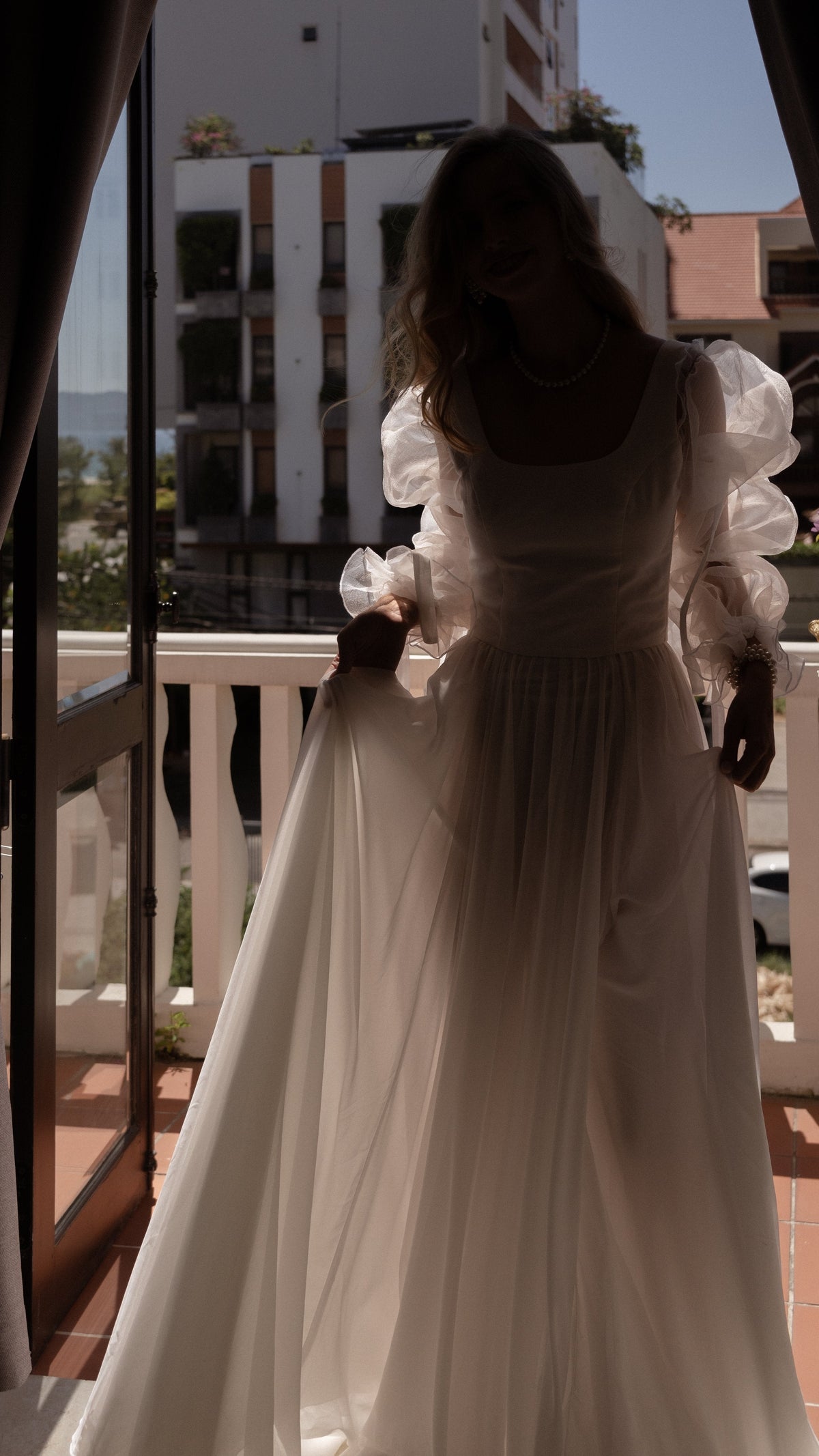 Bride silhouetted in a balcony doorway, light glowing through the sheer sleeves and chiffon skirt of a wedding dress, LUNA
