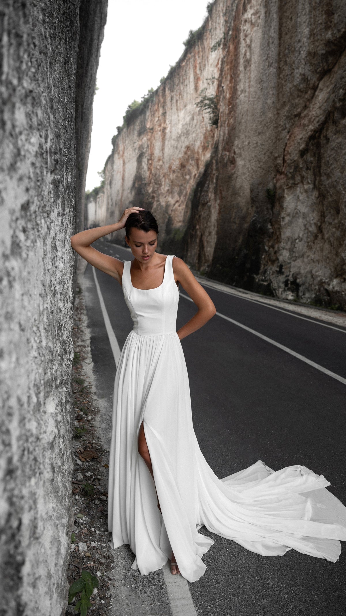 Sleeveless square-neck chiffon wedding dress with a side slit and long train, bride against a canyon wall on the road, LUNA