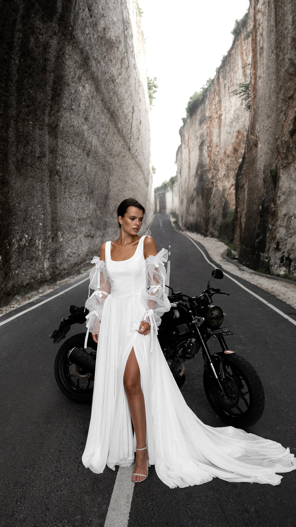 Front view on a canyon road of a minimalist chiffon wedding dress with off-shoulder organza sleeves and a thigh slit, motorbike behind, LUNA