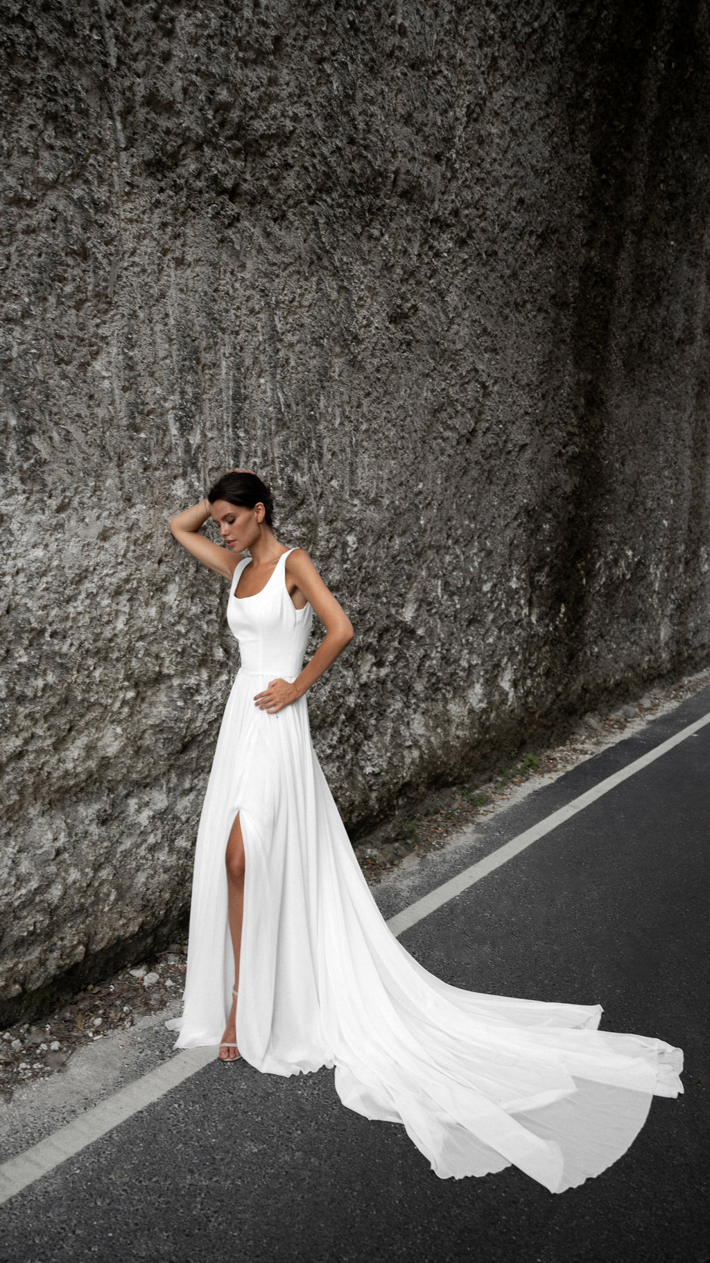Bride with a hand on her head under towering rock walls, sleeveless chiffon wedding dress with a train fanned on the asphalt, LUNA