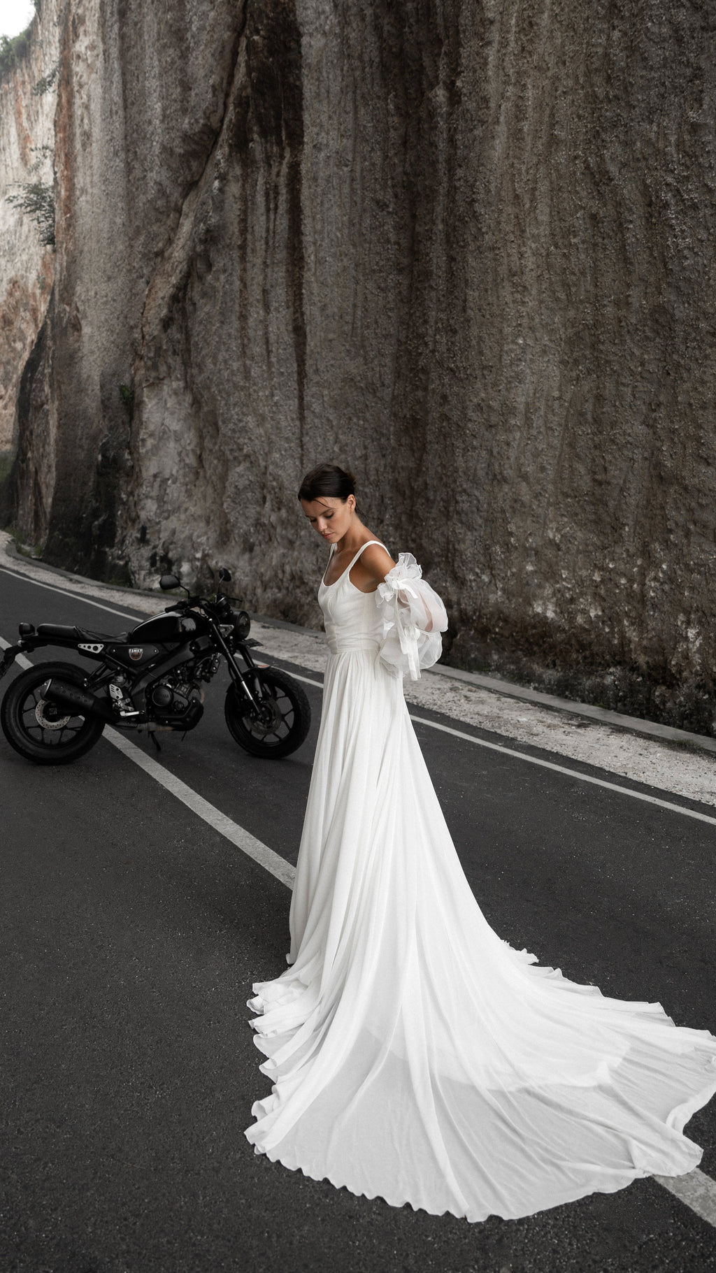 Bride walking along a canyon road, long chiffon train of a square-neck wedding dress trailing behind, motorbike parked, LUNA