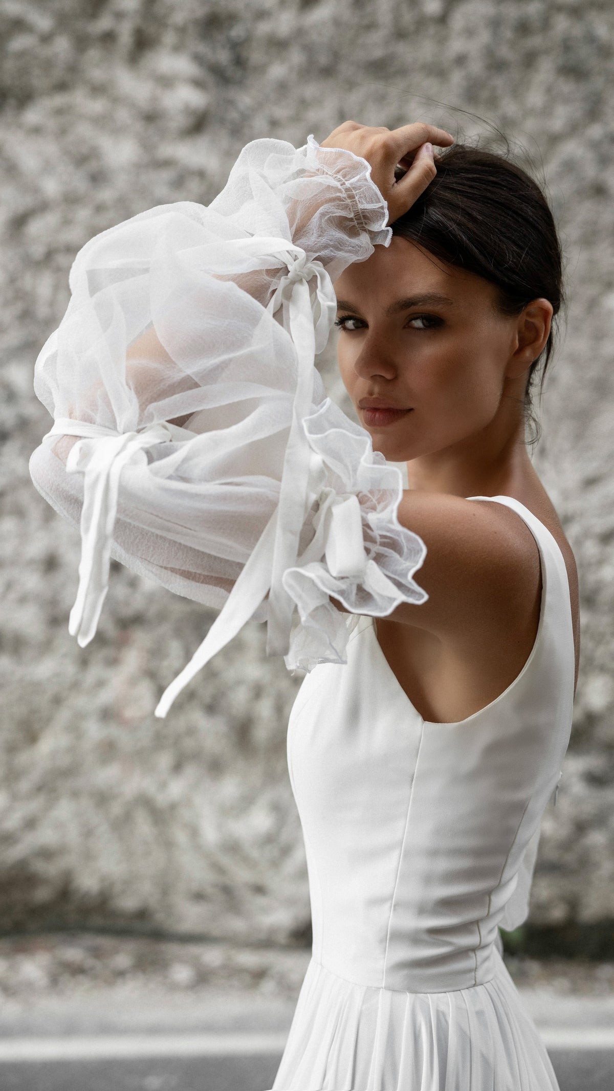 Close crop of an organza sleeve raised over the head, wide strap and fitted bodice of a chiffon wedding dress, rock wall behind, LUNA