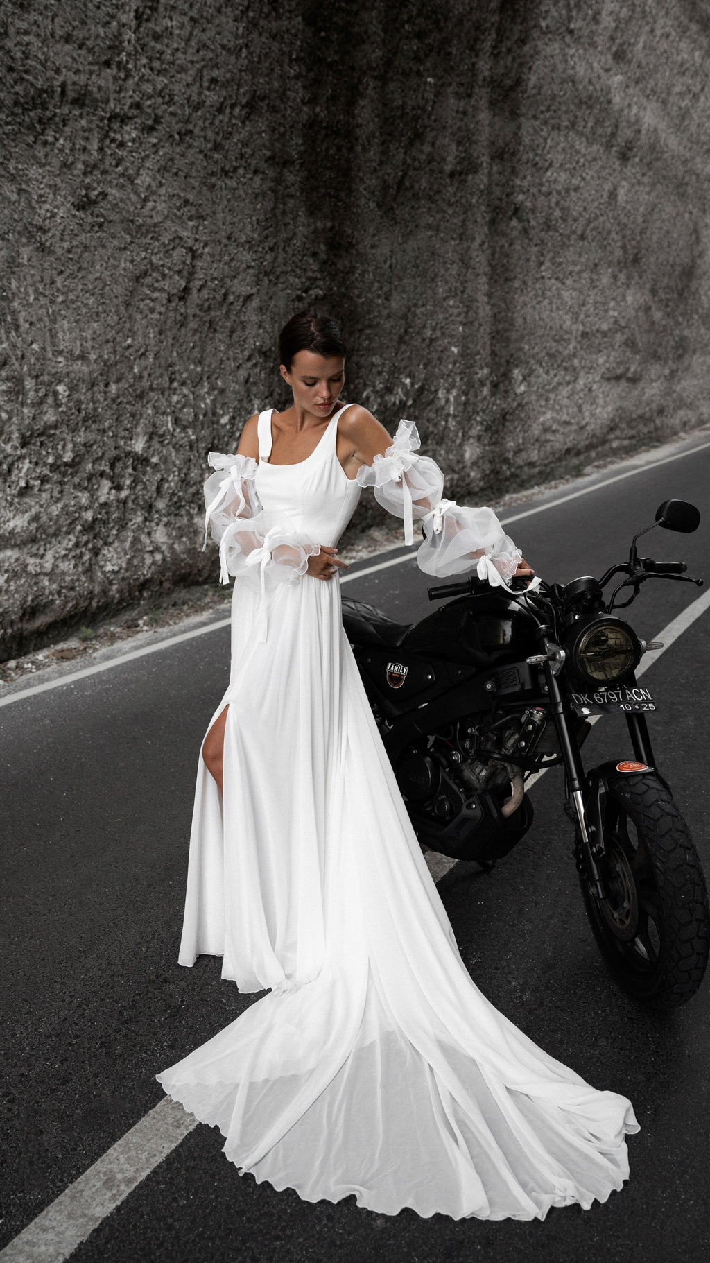 Bride resting a hand on a motorbike, chiffon wedding dress with off-shoulder sleeves and a train spread across the lane line, LUNA