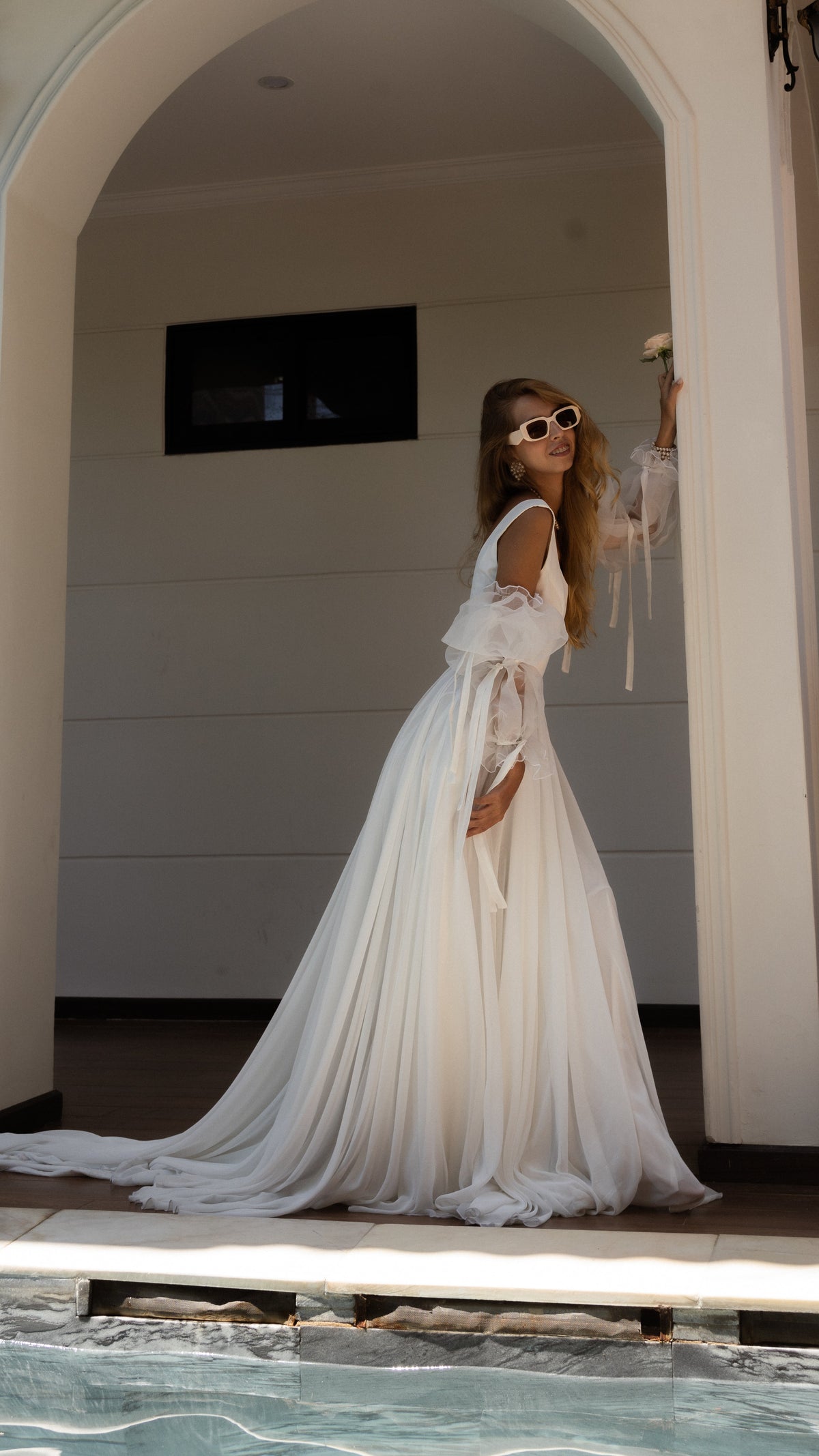 Bride in sunglasses posing in a white arch by a pool, chiffon wedding dress with puff organza sleeves and a sweeping train, LUNA