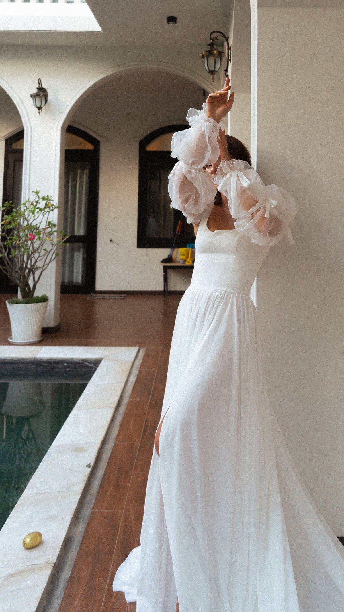 Bride stretching up against a pillar by a courtyard pool, square-neck wedding dress with off-shoulder organza sleeves, LUNA