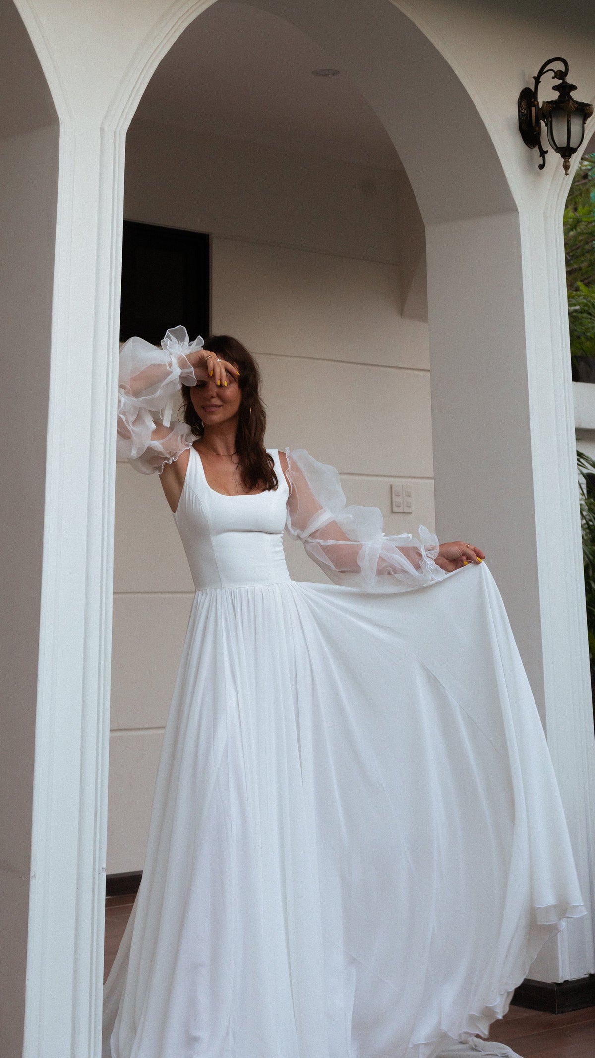 Bride shading her eyes in a white arch, holding out the chiffon skirt of a square-neck wedding dress with sheer sleeves, LUNA