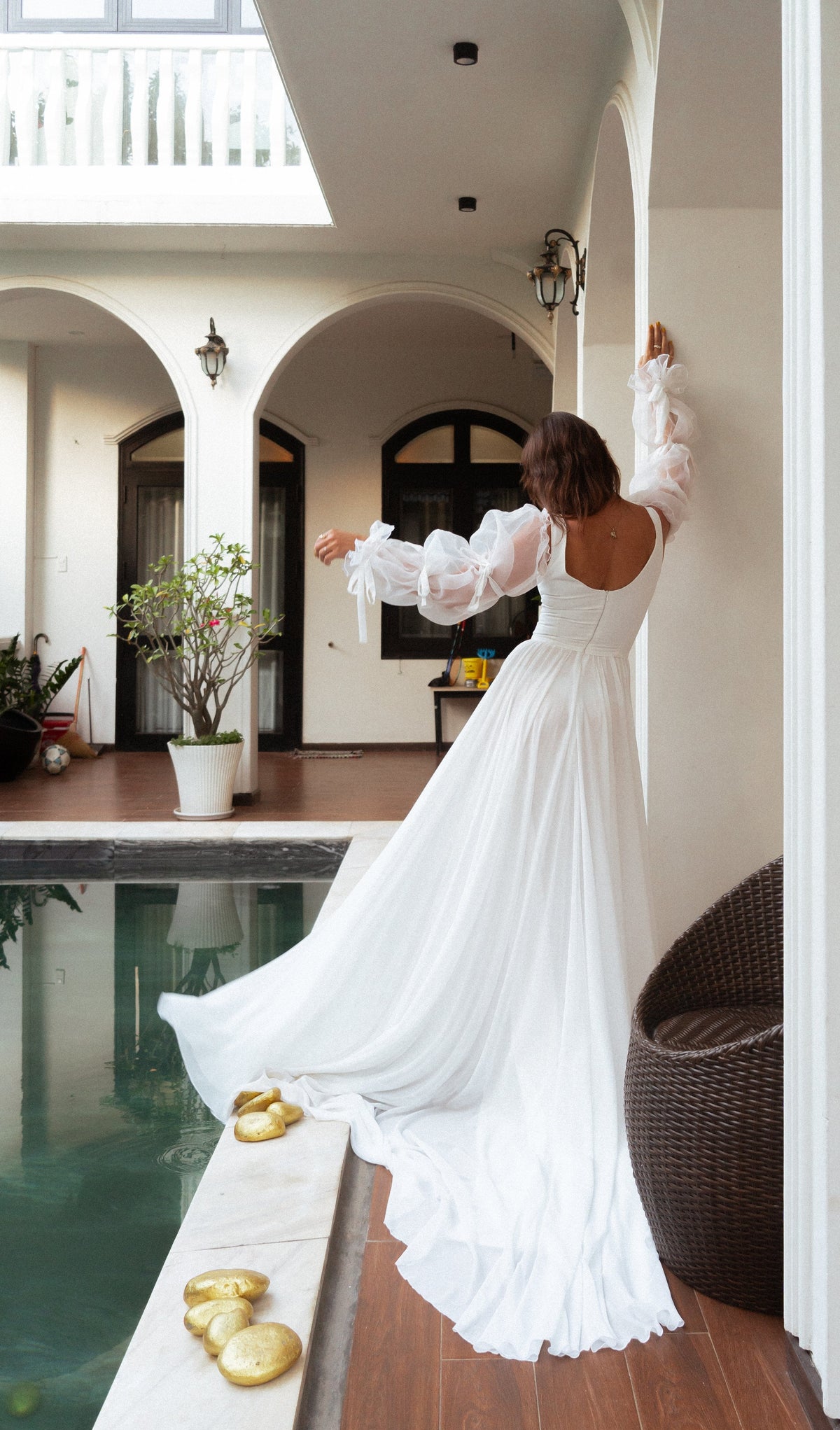 Back view of a scoop-back chiffon wedding dress with ribboned organza sleeves and a long train by a courtyard pool, LUNA