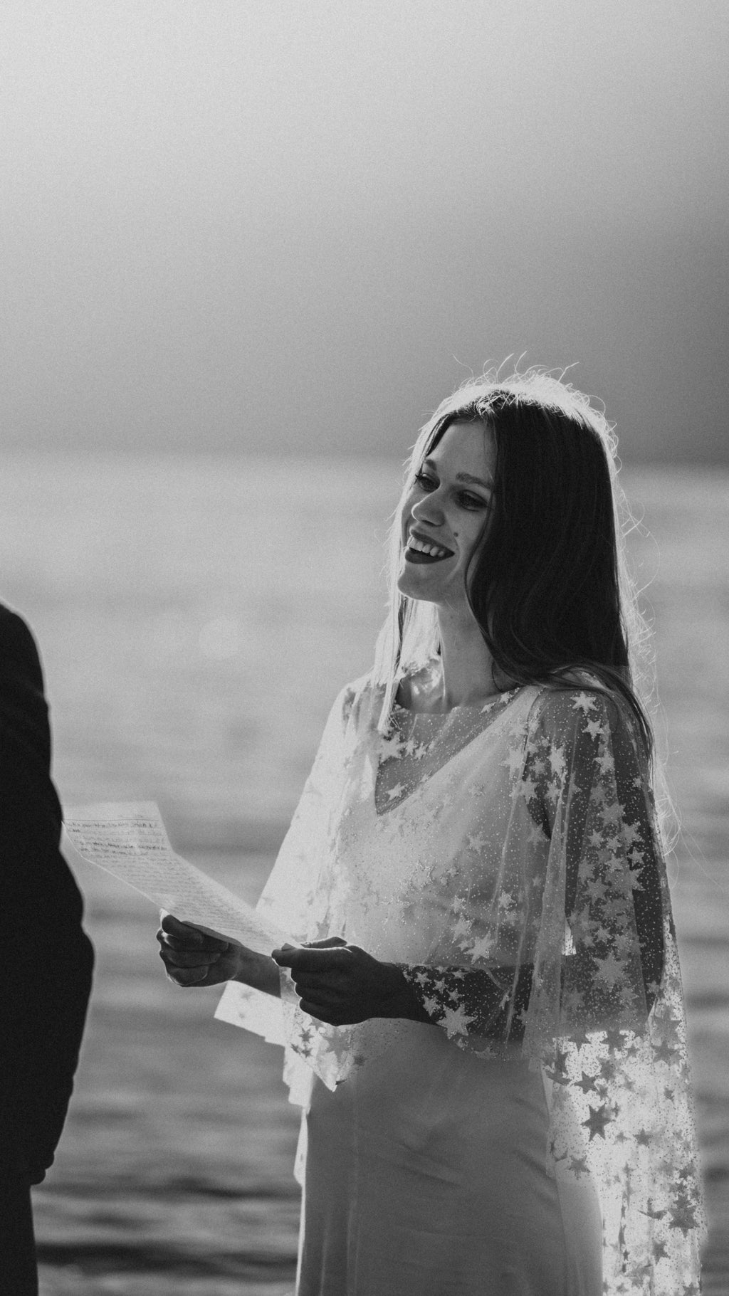Black and white photo of a smiling bride reading vows by a lake, star tulle bridal cape over her shoulders, Lunar Eclipse Cape Ivory