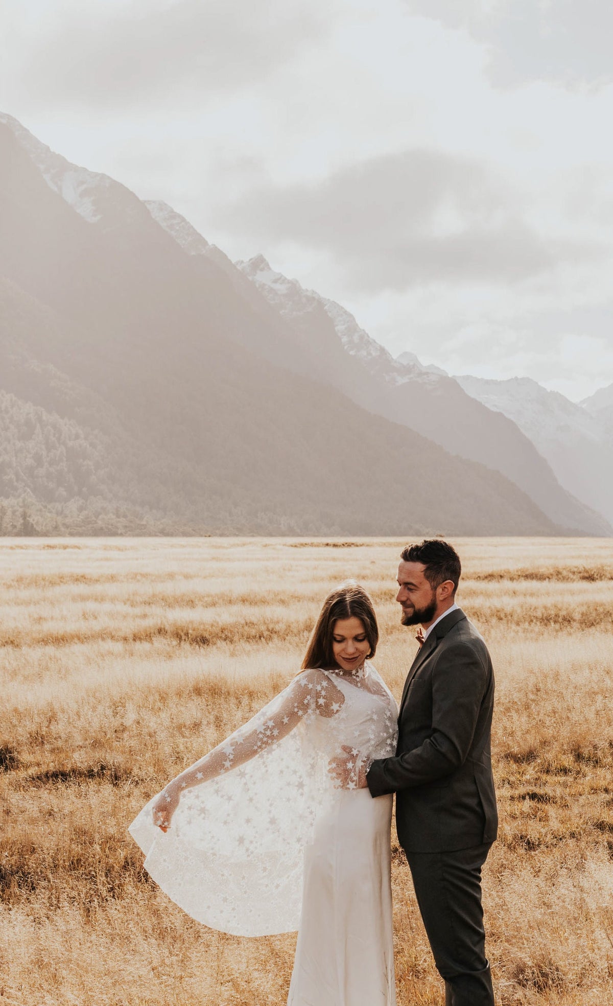Elopement couple in a golden grass valley under mountains, bride holding out the edge of a star bridal cape, Lunar Eclipse Cape Ivory