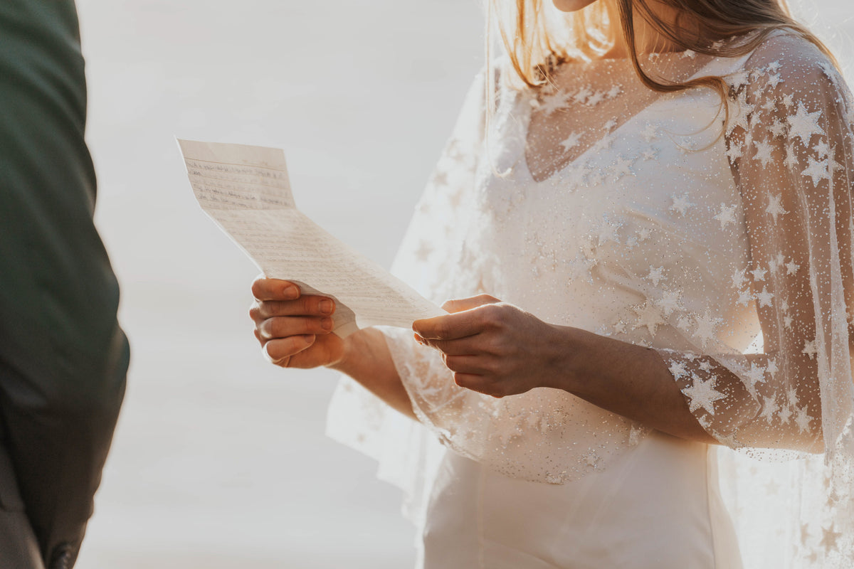 Close crop of hands holding vow notes, sheer ivory bridal cape with scattered stars over a simple gown, Lunar Eclipse Cape Ivory