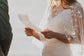 Close crop of hands holding vow notes, sheer ivory bridal cape with scattered stars over a simple gown, Lunar Eclipse Cape Ivory