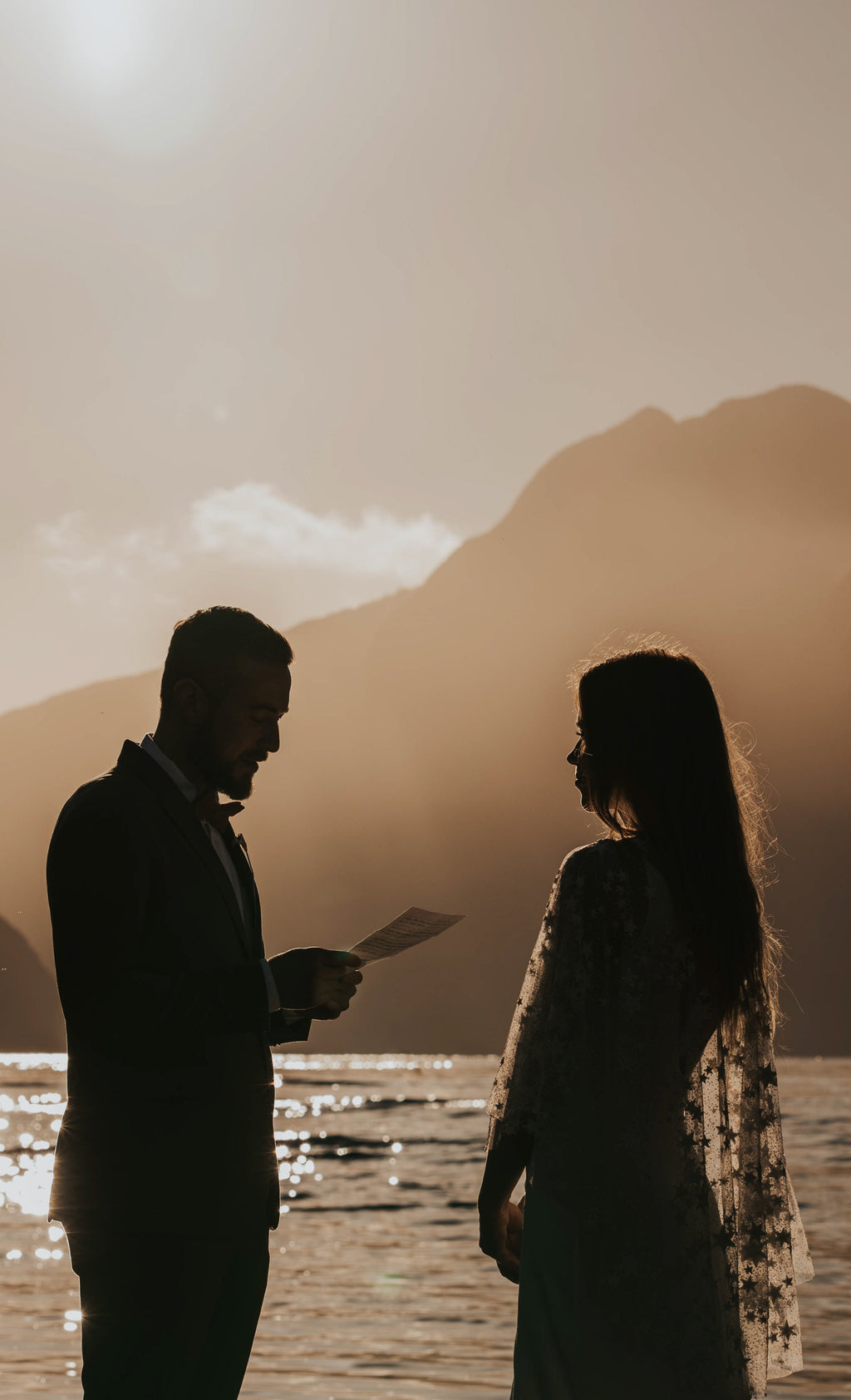 Silhouettes of a couple exchanging vows at a sunlit lake, stars visible along the edge of the bridal cape, Lunar Eclipse Cape Ivory