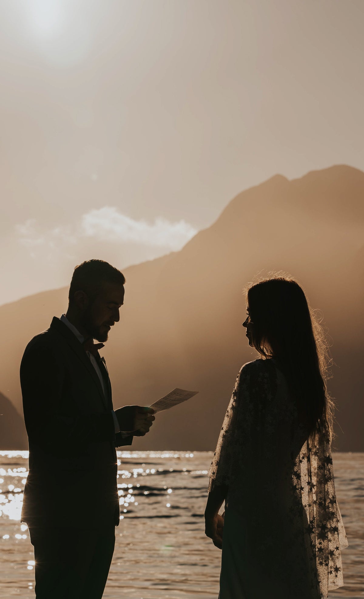 Silhouettes of a couple exchanging vows at a sunlit lake, stars visible along the edge of the bridal cape, Lunar Eclipse Cape Ivory