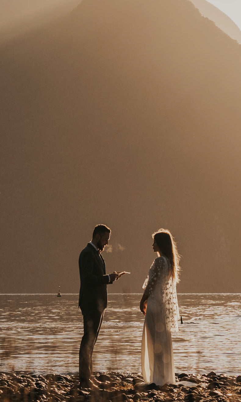 Couple facing each other on a stony lakeshore below a misty peak, bride in a star bridal cape over a long gown, Lunar Eclipse Cape Ivory