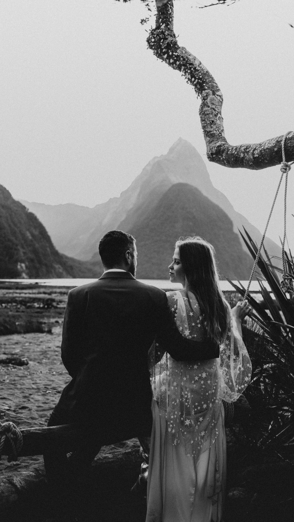 Black and white photo of a couple by the water below a sharp mountain peak, star tulle bridal cape over the bride, Lunar Eclipse Cape Ivory