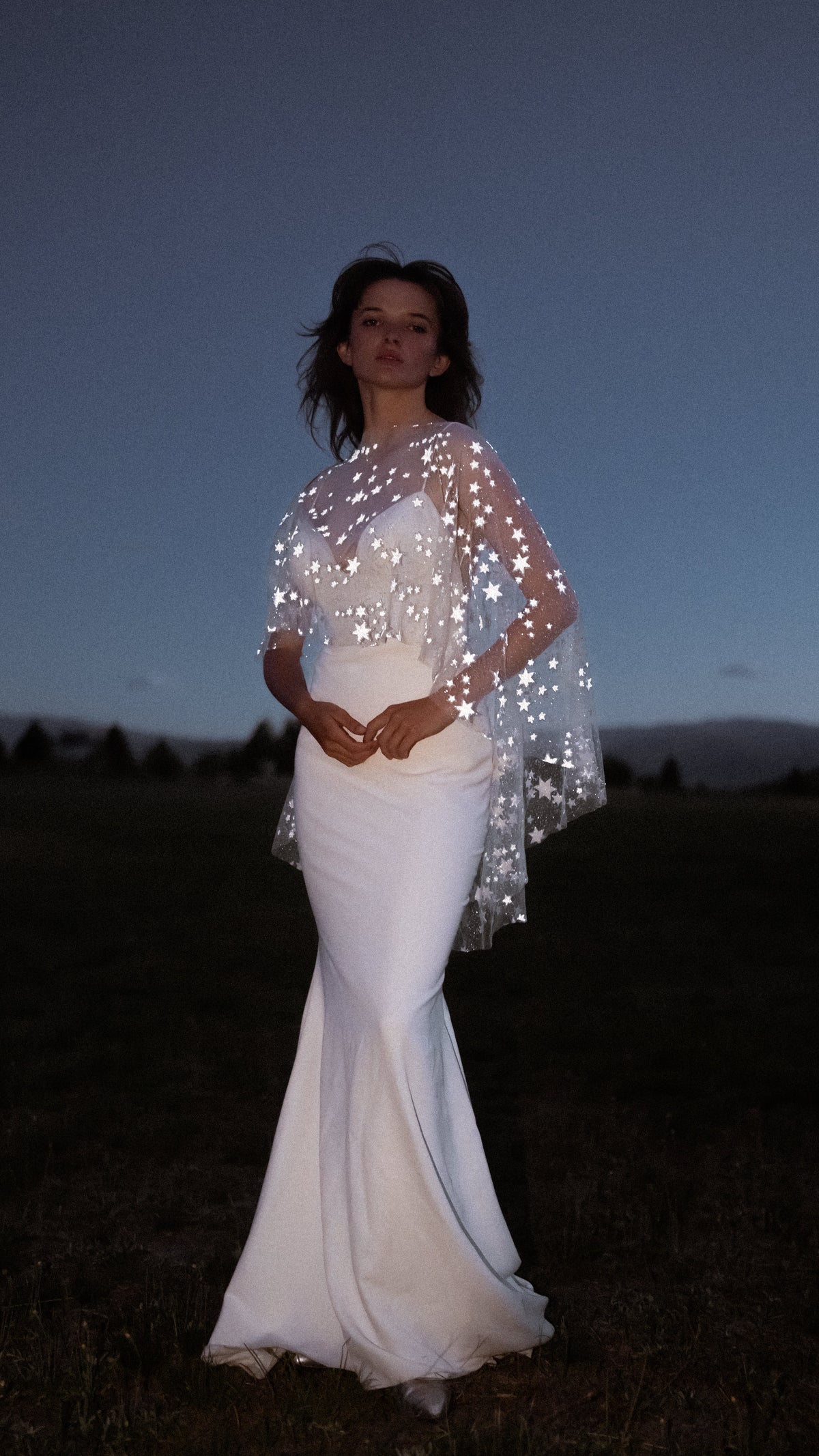 Bride in an evening field, hands clasped at the waist, star bridal cape shining over a strapless fitted gown, Lunar Eclipse Cape Ivory