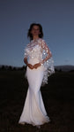 Bride in an evening field, hands clasped at the waist, star bridal cape shining over a strapless fitted gown, Lunar Eclipse Cape Ivory