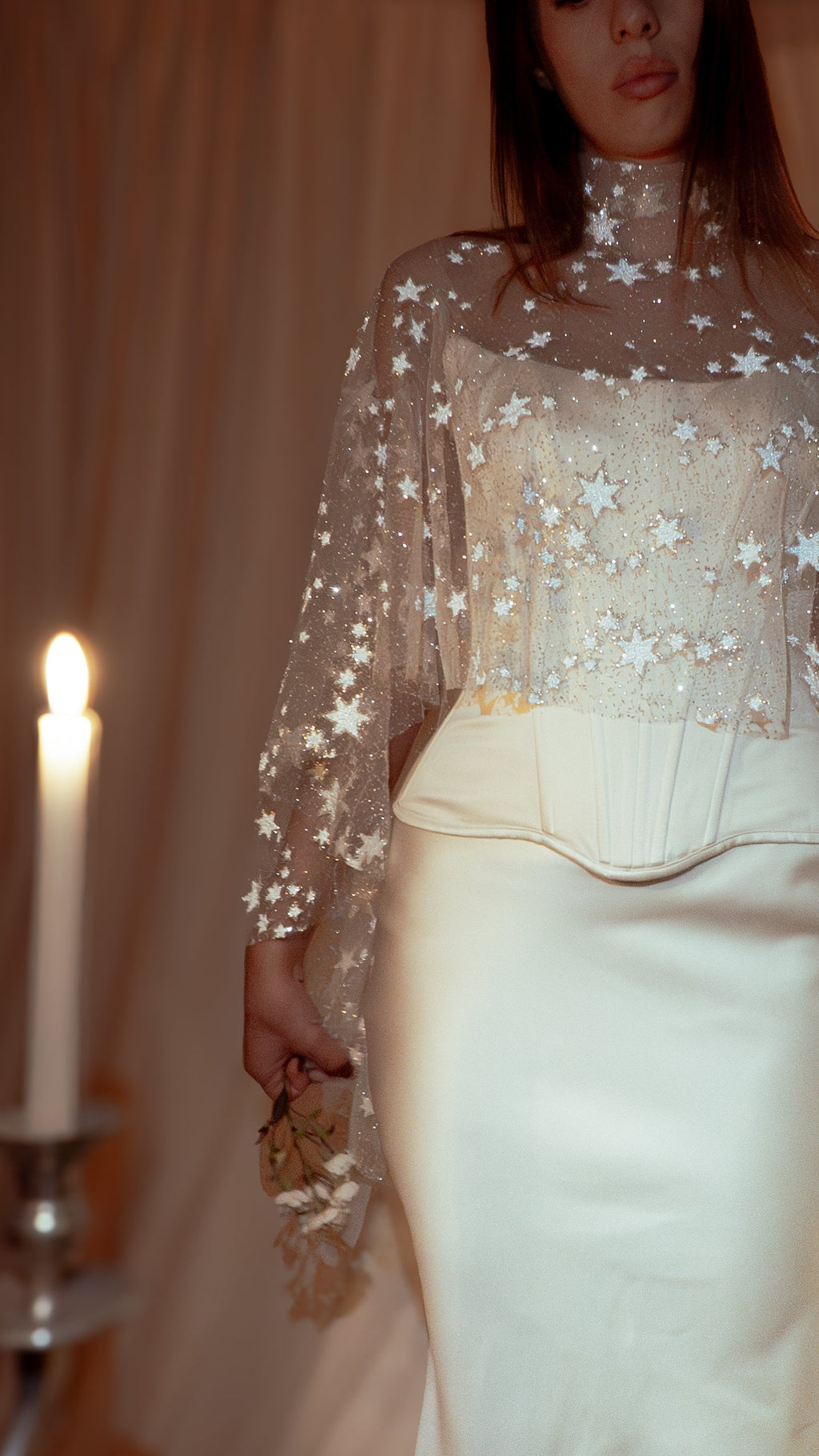 Close crop of a high-neck star tulle bridal cape over a corset bodice, bride holding dried flowers beside a candle, Lunar Eclipse Cape Ivory