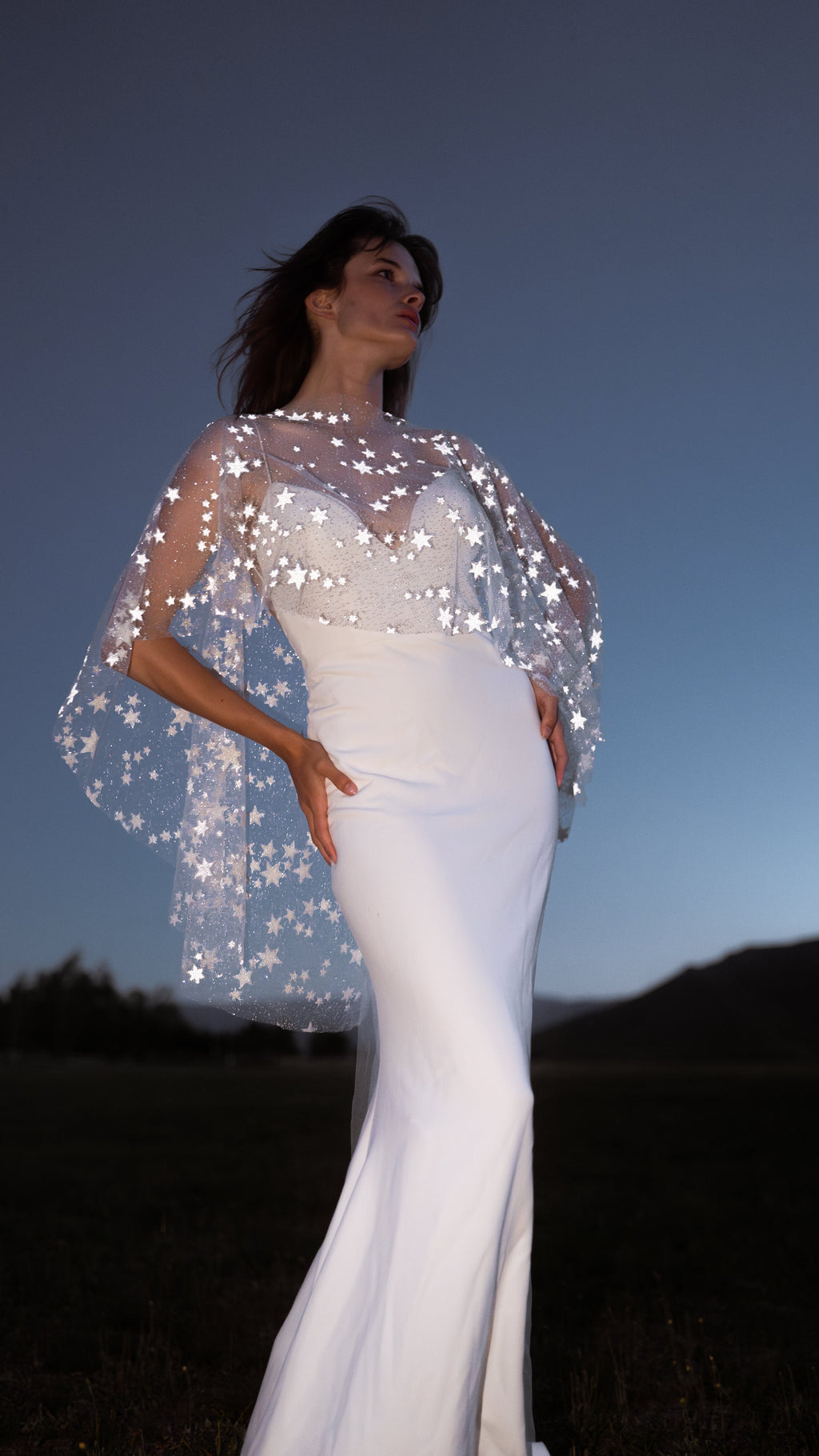 Bride at dusk on a dark field, reflective stars on a short ivory bridal cape catching the light over a fitted gown, Lunar Eclipse Cape Ivory
