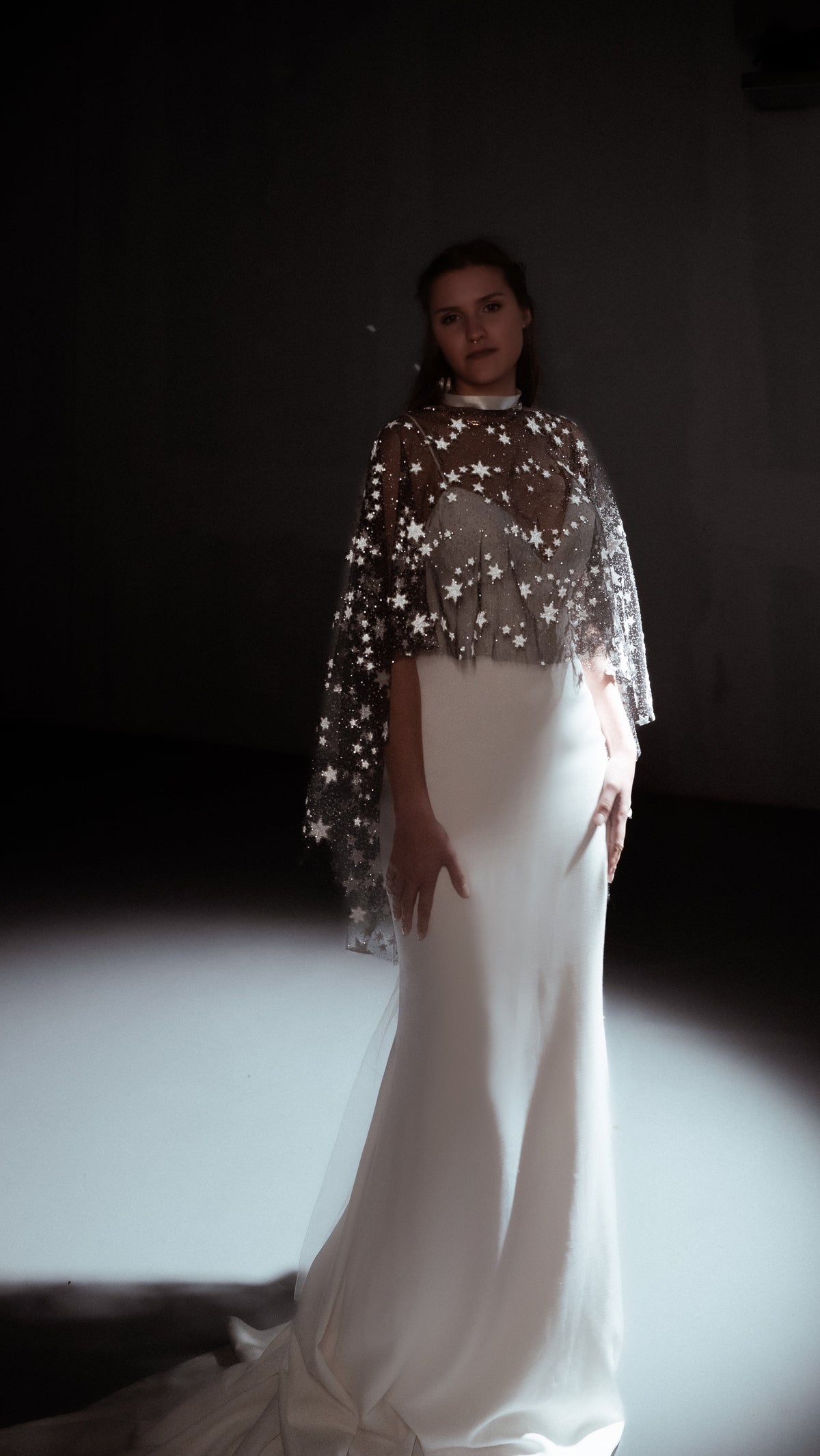 Full-length shot in a dark room, star bridal cape glinting in a pool of light over a column gown, Lunar Eclipse Cape Ivory