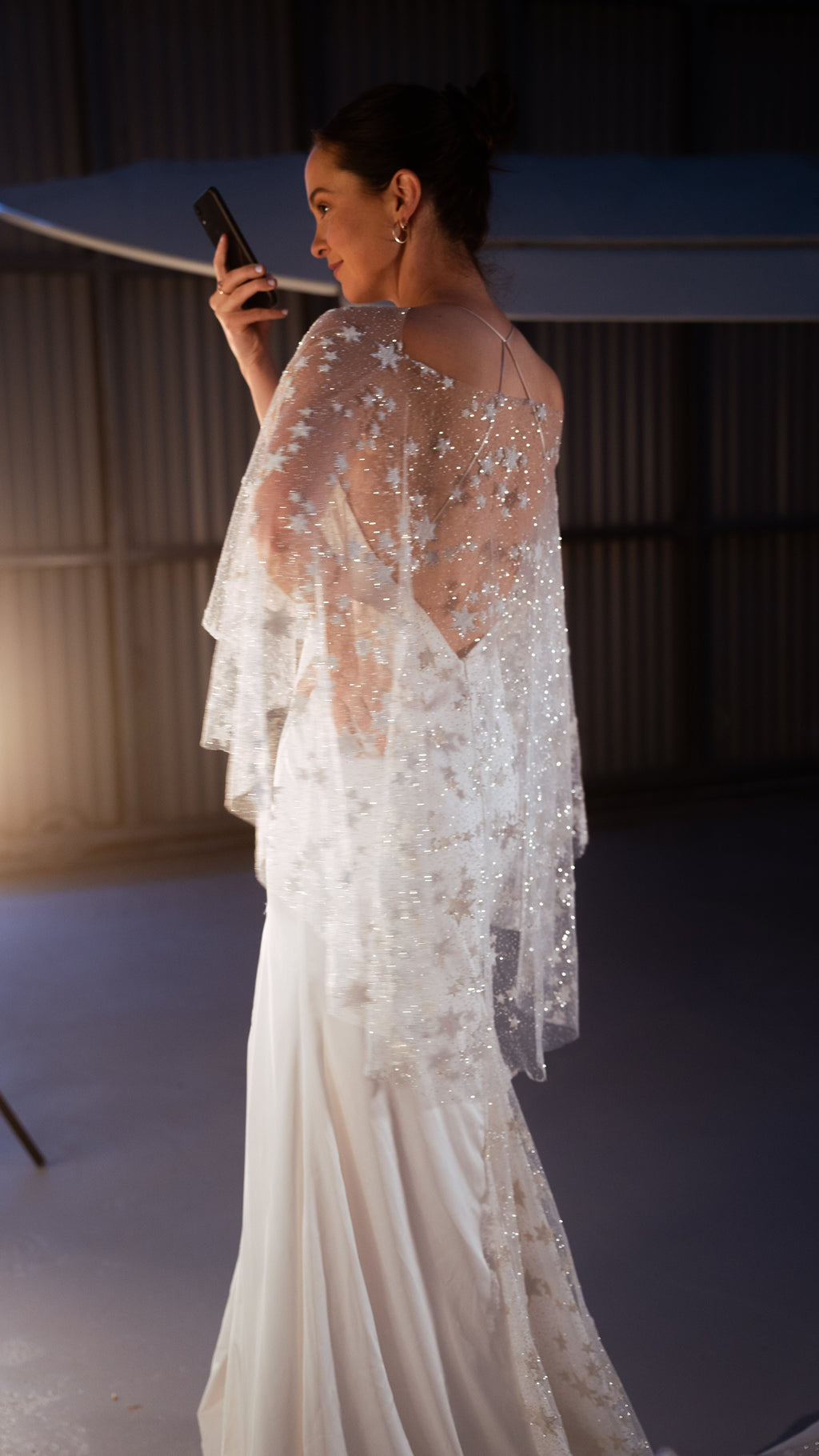 Bride filming herself on a phone, rear view of a sheer star bridal cape over an open-back slip gown in a hangar, Lunar Eclipse Cape Ivory