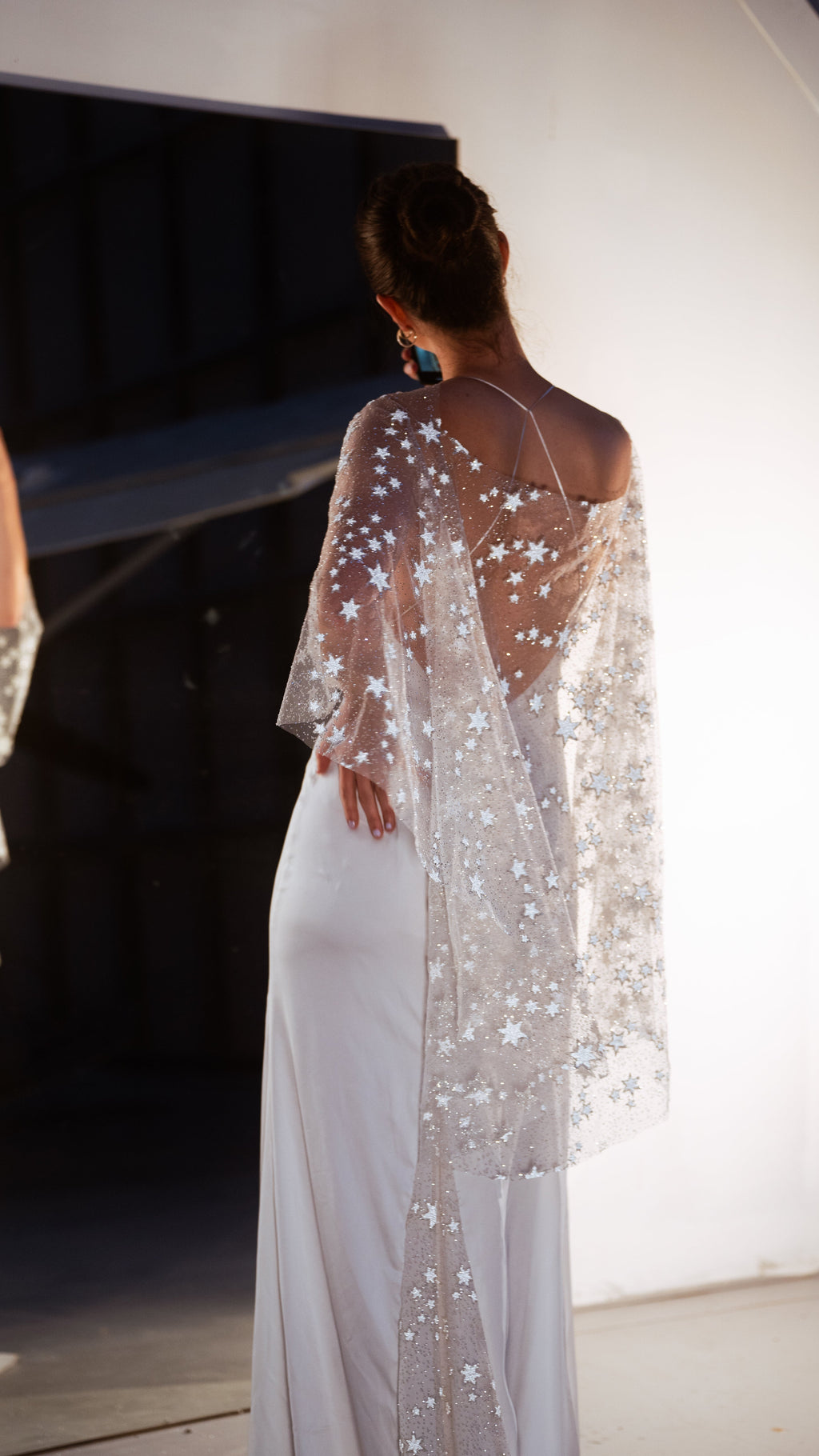 Back view of a bride with a hand on her hip, sheer star bridal cape flowing past the waist of a slim gown, Lunar Eclipse Cape Ivory