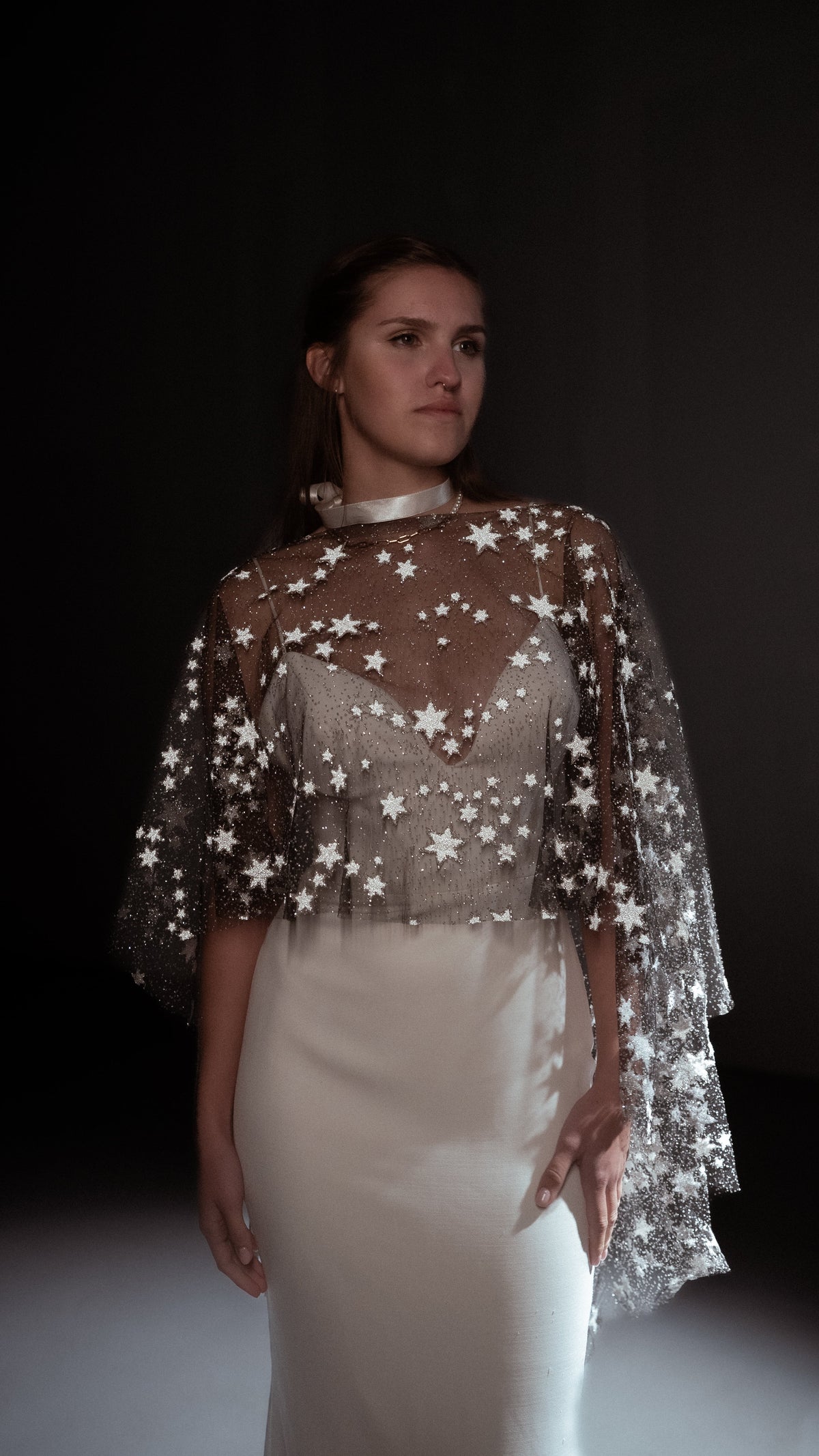 Black celestial bridal cape with white stars and a high neck over a satin gown and ribbon choker, dark studio. Lunar Eclipse Cape Black by Ephemeris