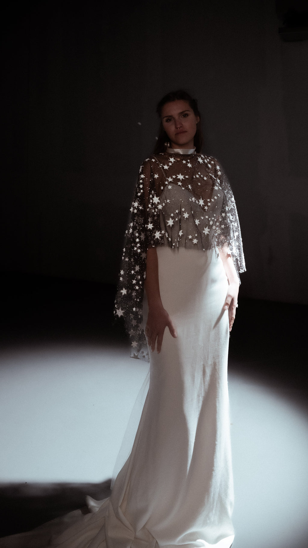 Full-length view of a black star tulle bridal cape for an alt bride, worn over an ivory mermaid gown with a train in a spotlight, Lunar Eclipse Cape Black
