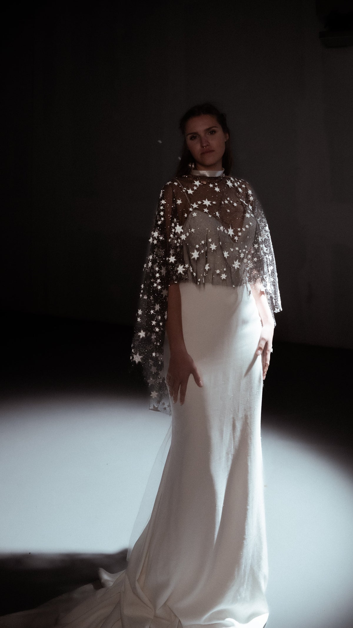 Full-length view of a black star tulle bridal cape for an alt bride, worn over an ivory mermaid gown with a train in a spotlight, Lunar Eclipse Cape Black