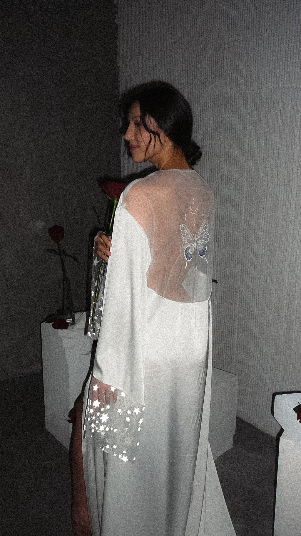 Bride glancing back holding a rose, butterfly on the sheer back of a long satin bridal robe, star cuff at her hip, Metamorphosis Robe