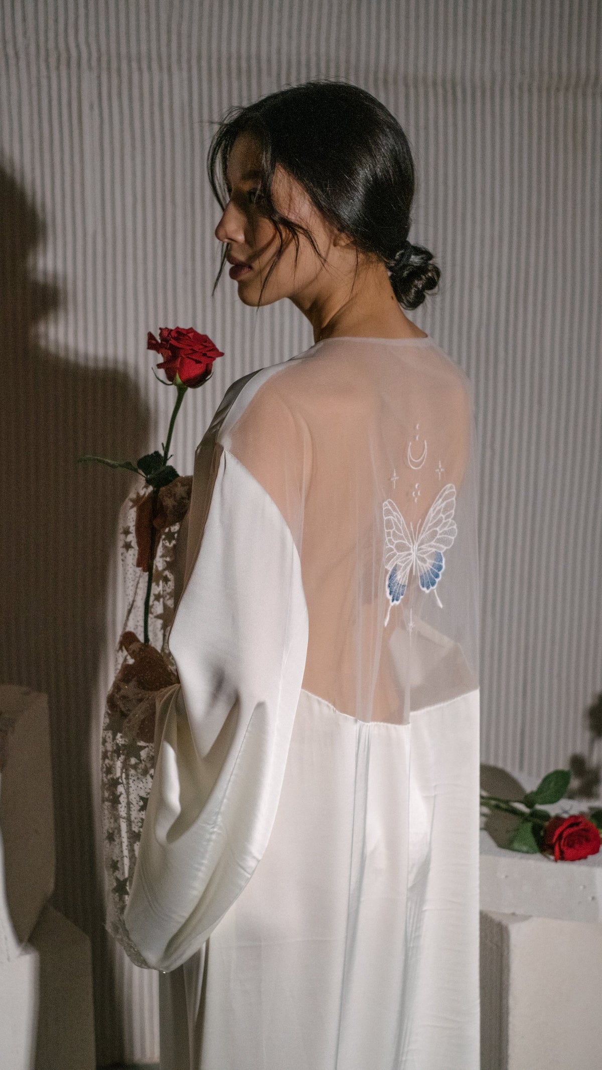 Bride looking over her shoulder holding a red rose, sheer back and embroidered butterfly of an ivory bridal robe, Metamorphosis Robe