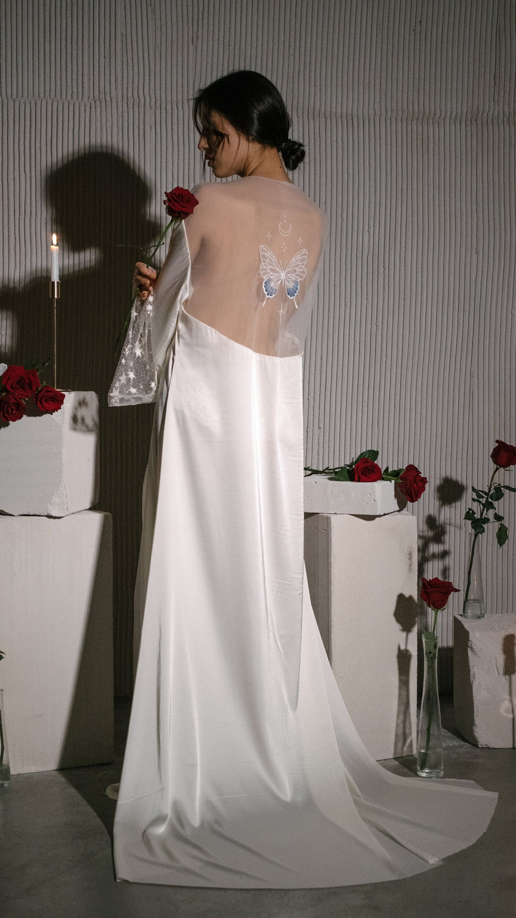 Rear view of a long satin bridal robe with a sheer butterfly back panel, rose in hand, candles and plinths, Metamorphosis Robe