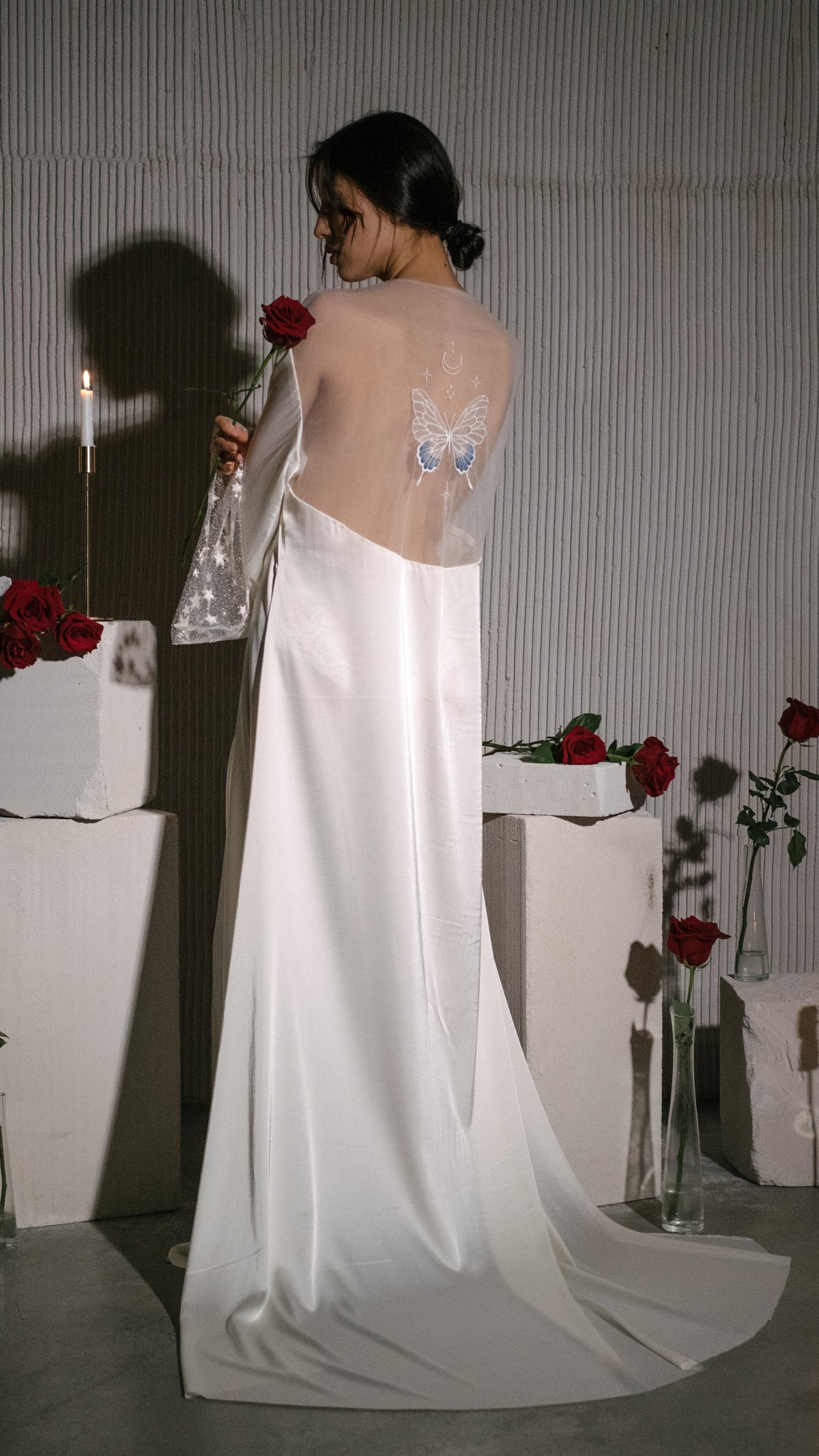 Rear view of a long satin bridal robe with a sheer butterfly back panel, rose in hand, candles and plinths, Metamorphosis Robe