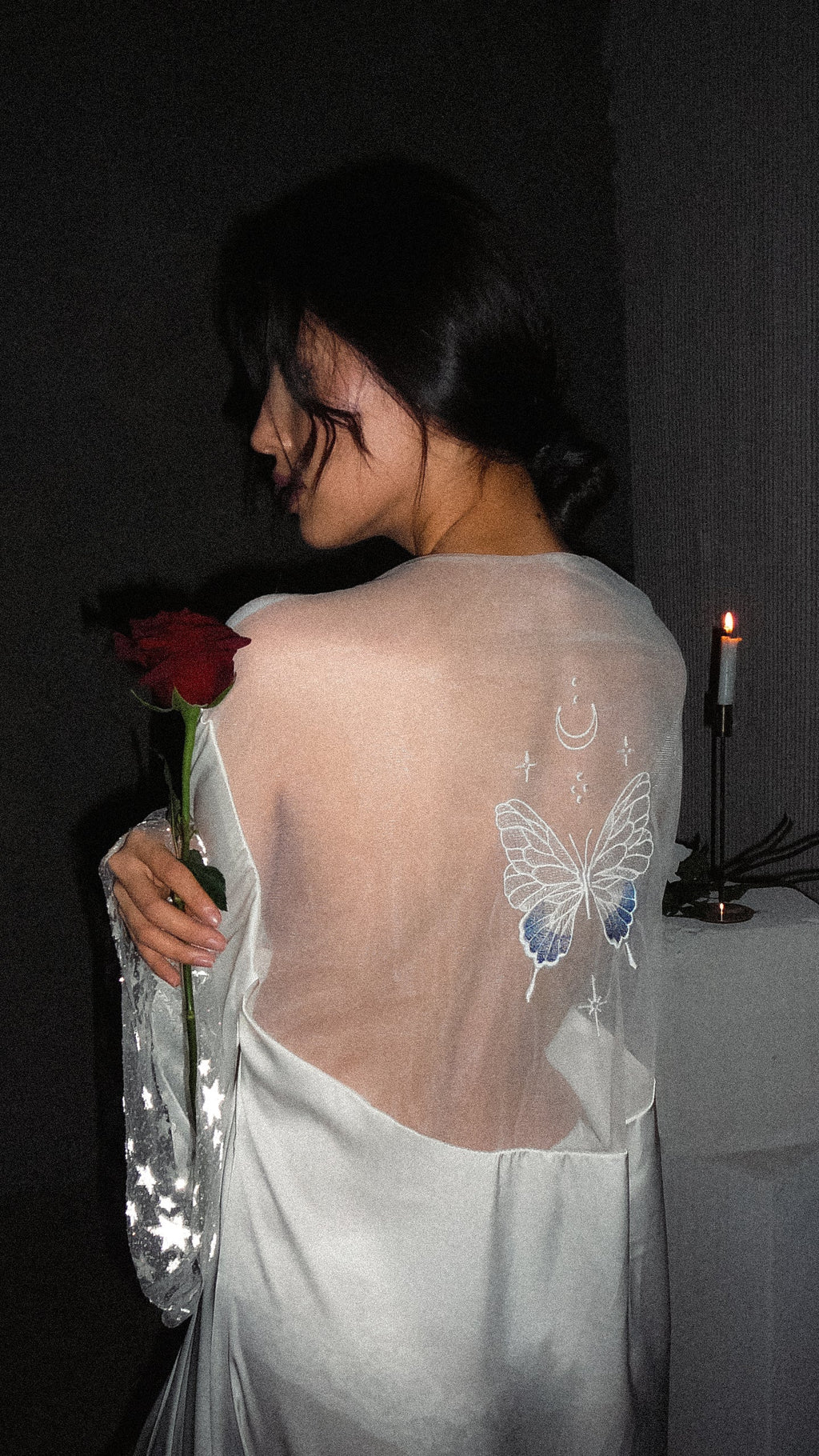 Over-the-shoulder view of a sheer bridal robe back with an ivory and blue butterfly, moon and stars, red rose in hand, Metamorphosis Robe