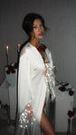 Getting-ready bridal robe in ivory satin with sheer star cuffs, bride holding a rose by candles and white plinths, Metamorphosis Robe