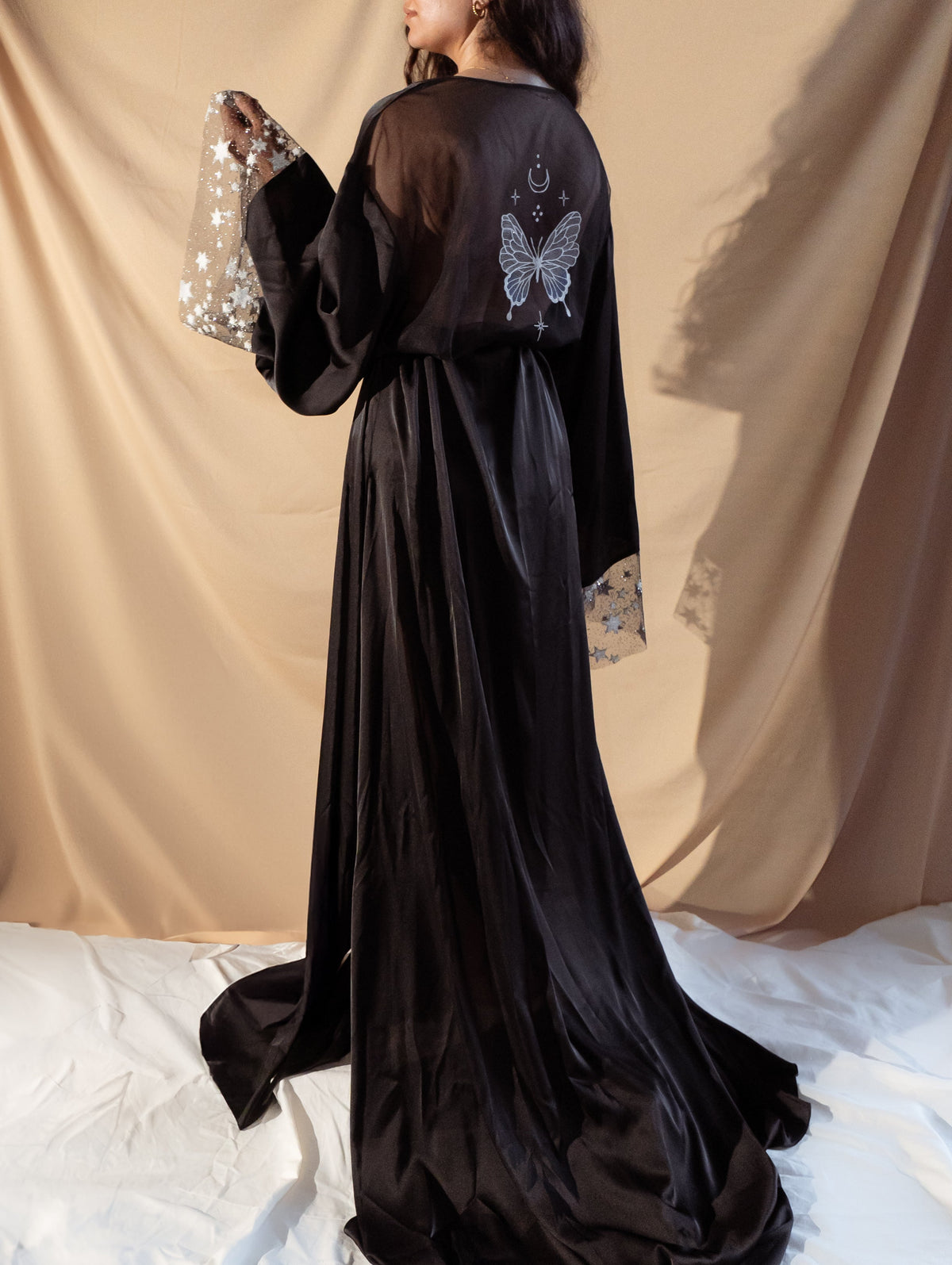 Back view of a black bridal robe with a long train, butterfly embroidery and starry sleeves, bride standing on white sheets, Metamorphosis Robe Black