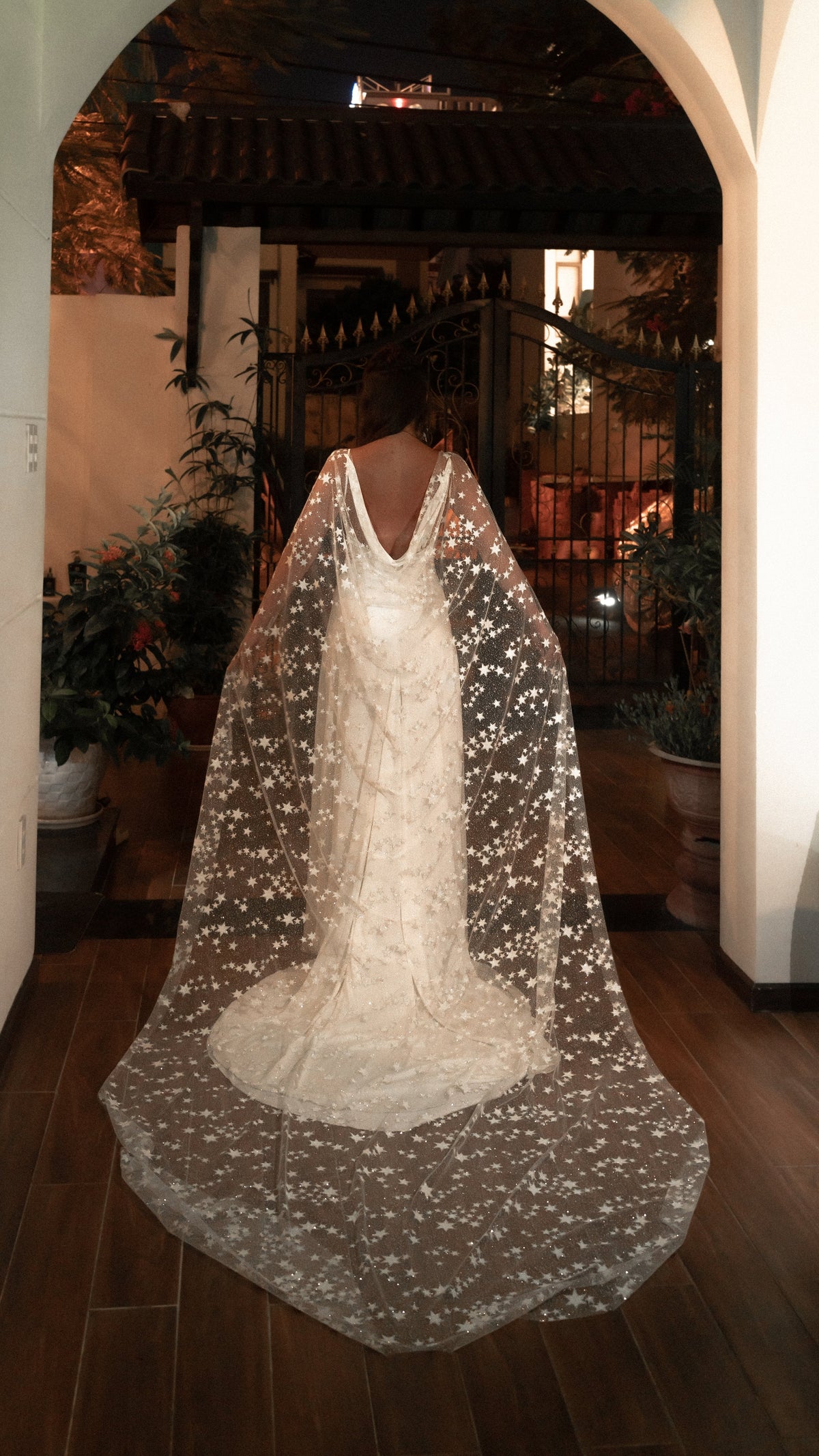 Bridal cape with scattered stars over a backless gown, bride walking toward a lit archway in the evening, Milky Way Cape Black