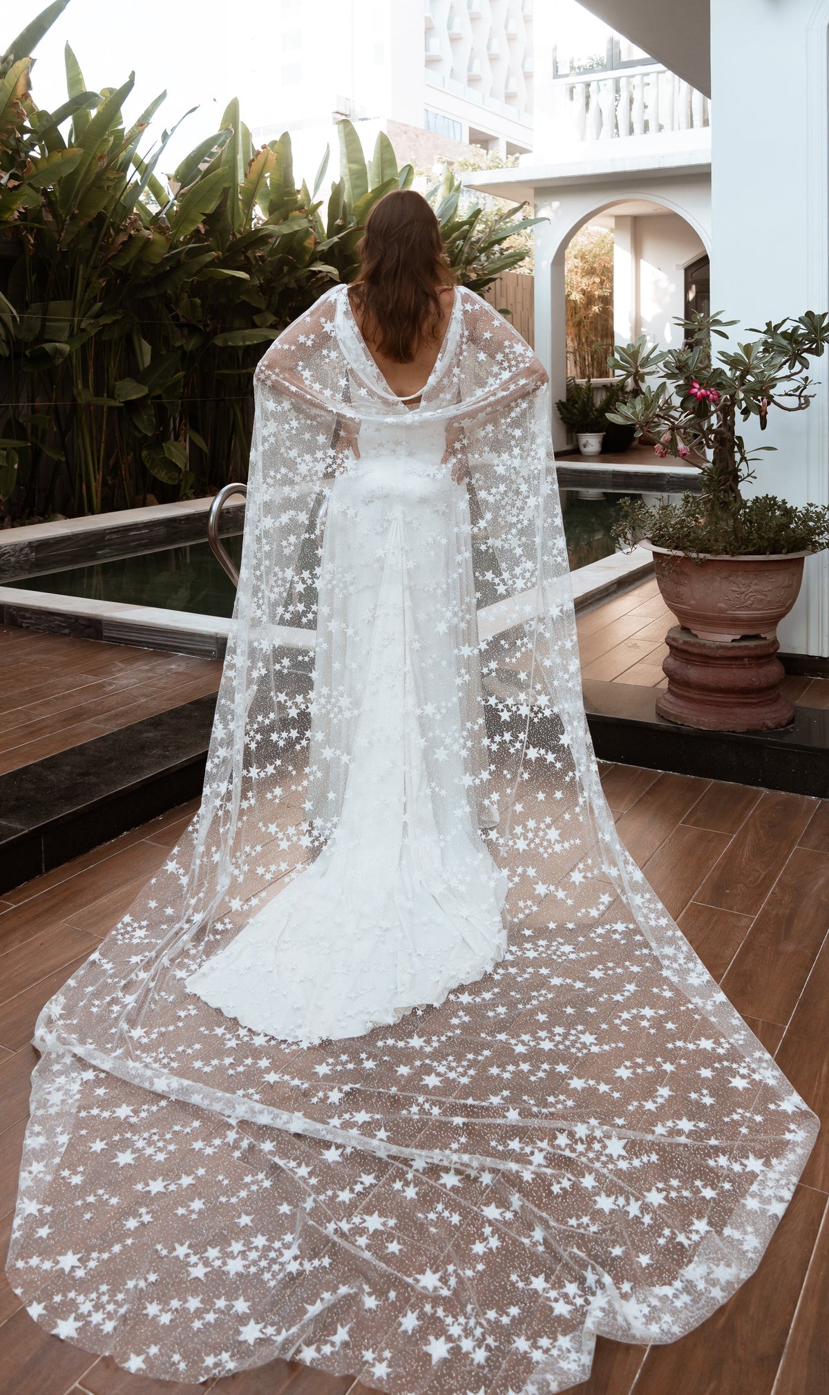 Star tulle bridal cape spread in a wide train across a poolside deck, bride seen from behind in daylight, Milky Way Cape
