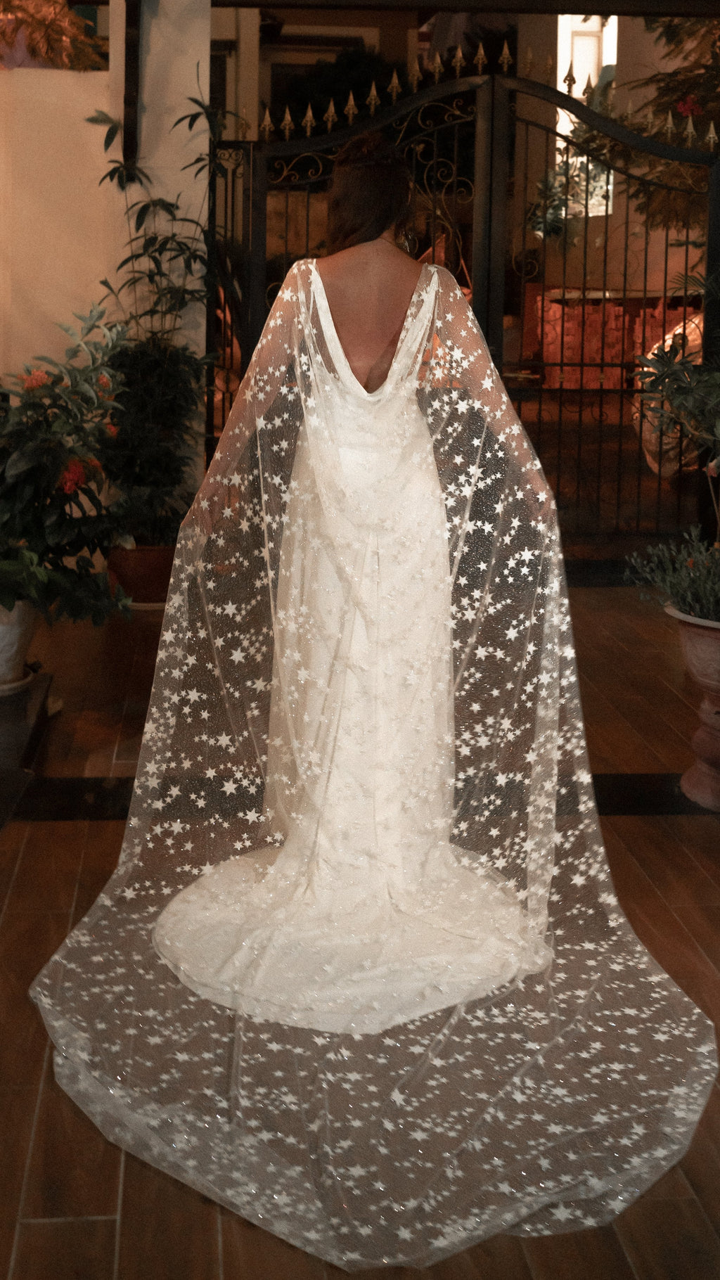 Back view of a bride in a star-scattered bridal cape and low-back ivory gown at an evening garden gate, Milky Way Cape