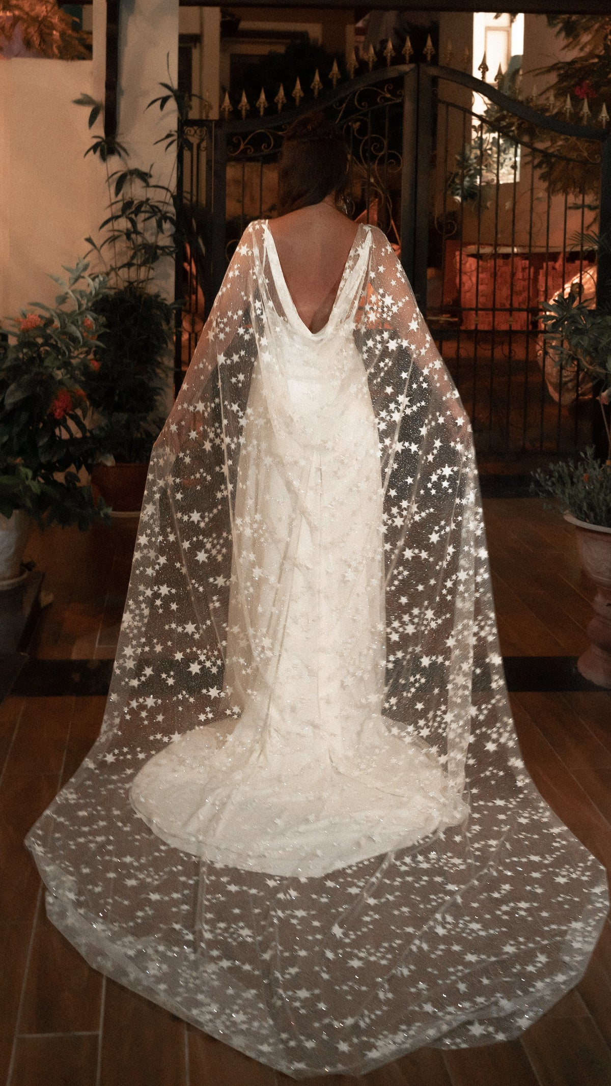 Back view of a bride in a star-scattered bridal cape and low-back ivory gown at an evening garden gate, Milky Way Cape