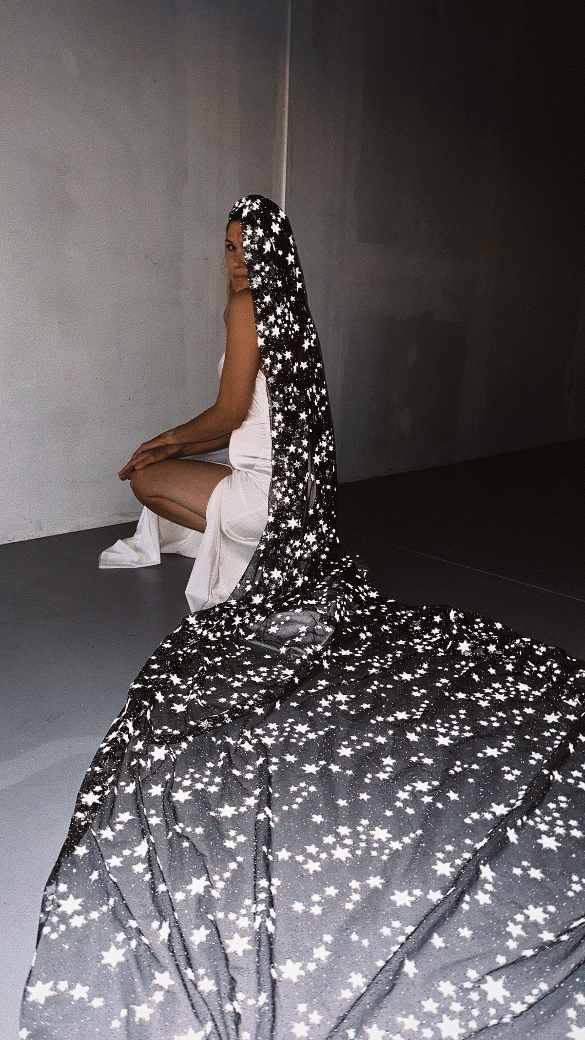 Long star wedding veil spread across a concrete floor, bride crouching in a white slip dress behind it, Milky Way Veil