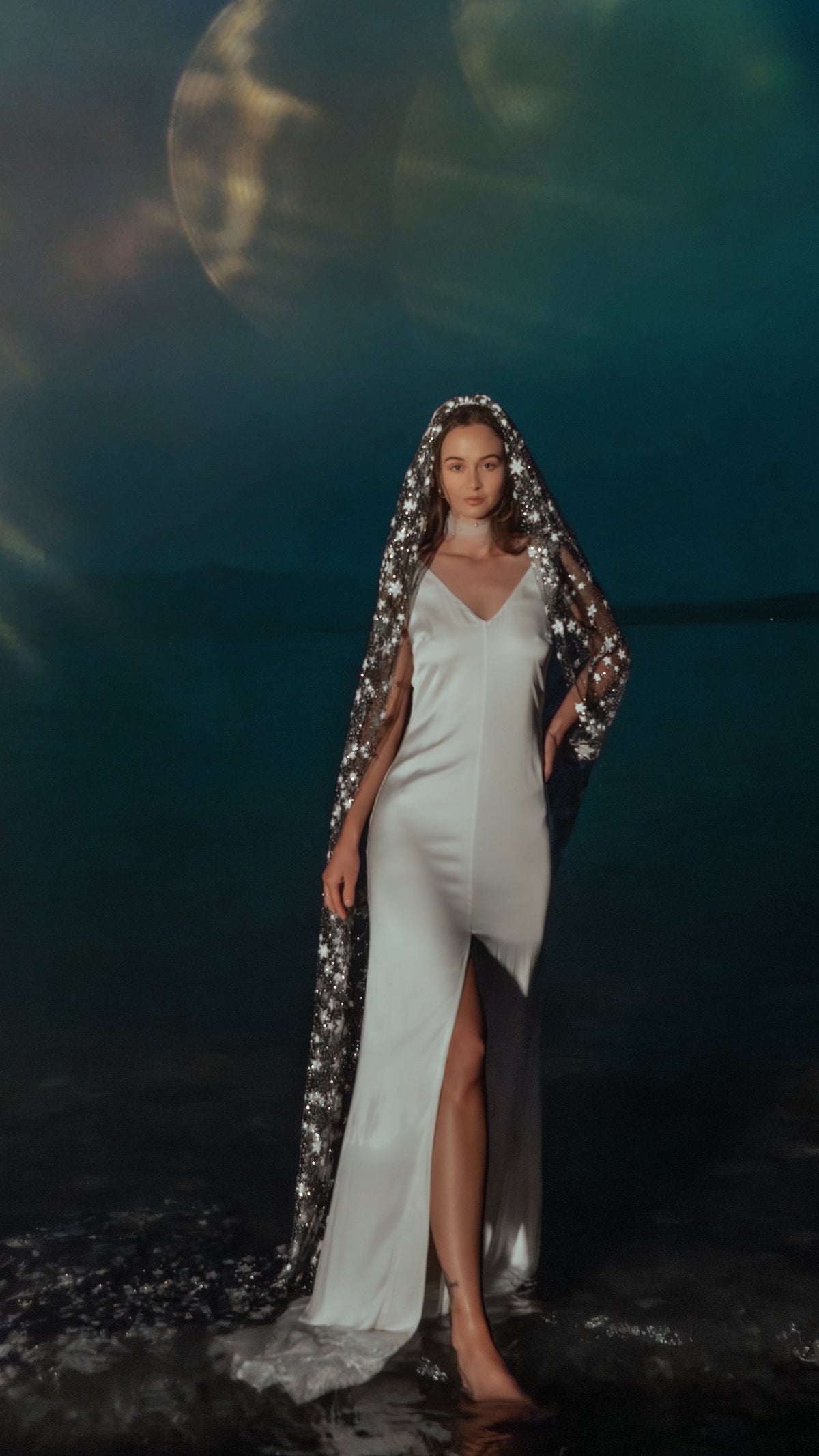 Bride in a V-neck slip dress standing in shallow water under a star wedding veil, large moon behind her, Milky Way Veil