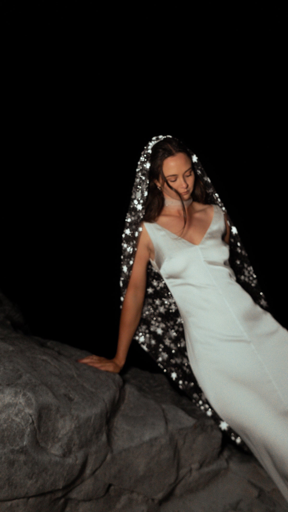 Bride leaning on a dark rock at night, star wedding veil framing her satin slip dress, Milky Way Veil