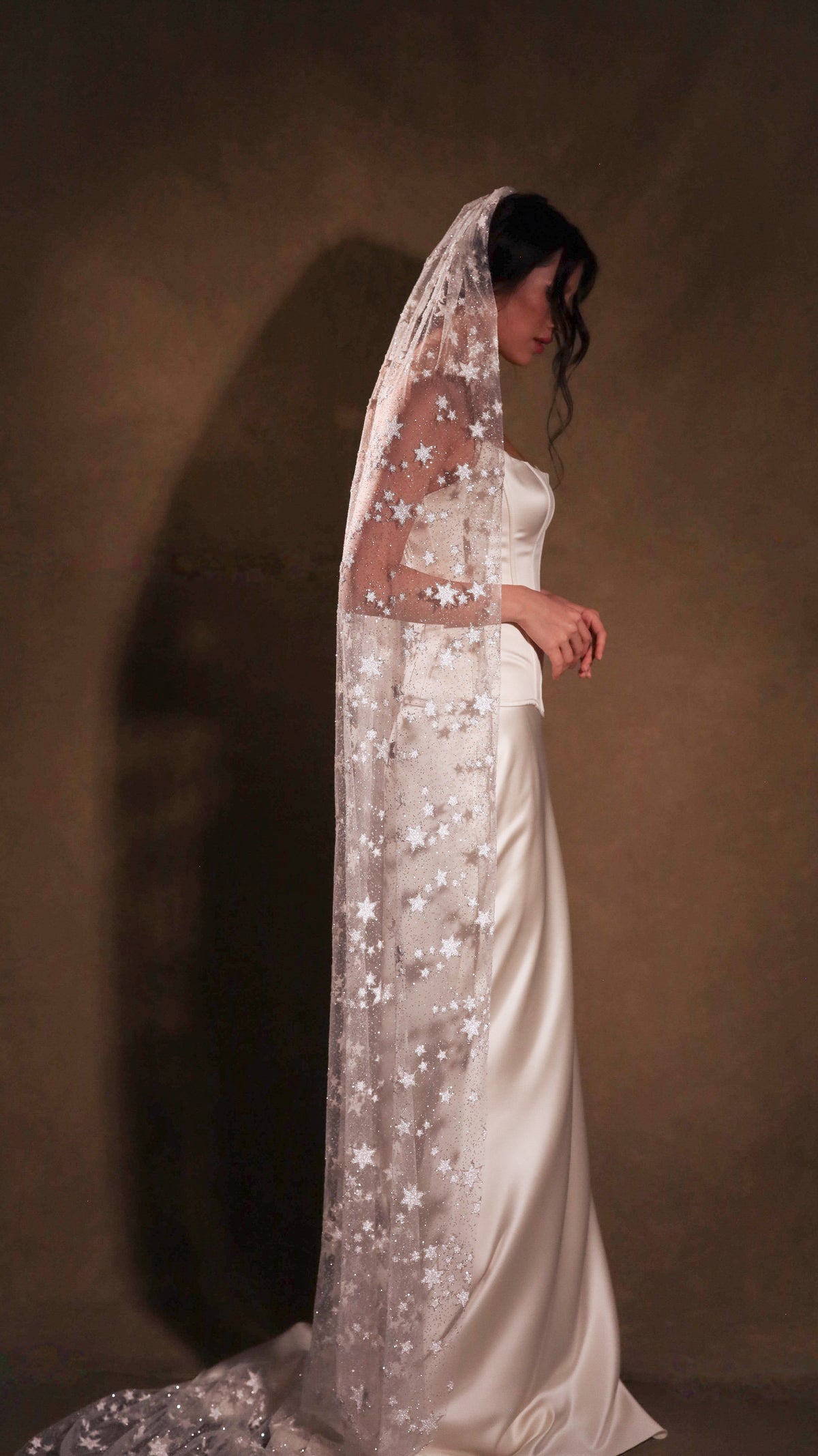 Bride with head bowed under a star wedding veil, side view in an ivory satin gown against a warm brown backdrop, Milky Way Veil