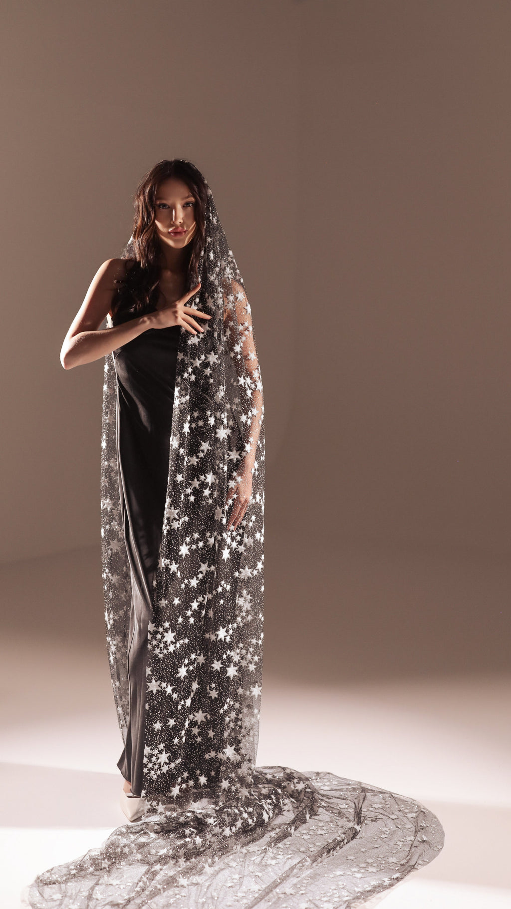 Model in a black slip dress under a long star wedding veil, hand at her chest, spotlit studio floor, Milky Way Veil
