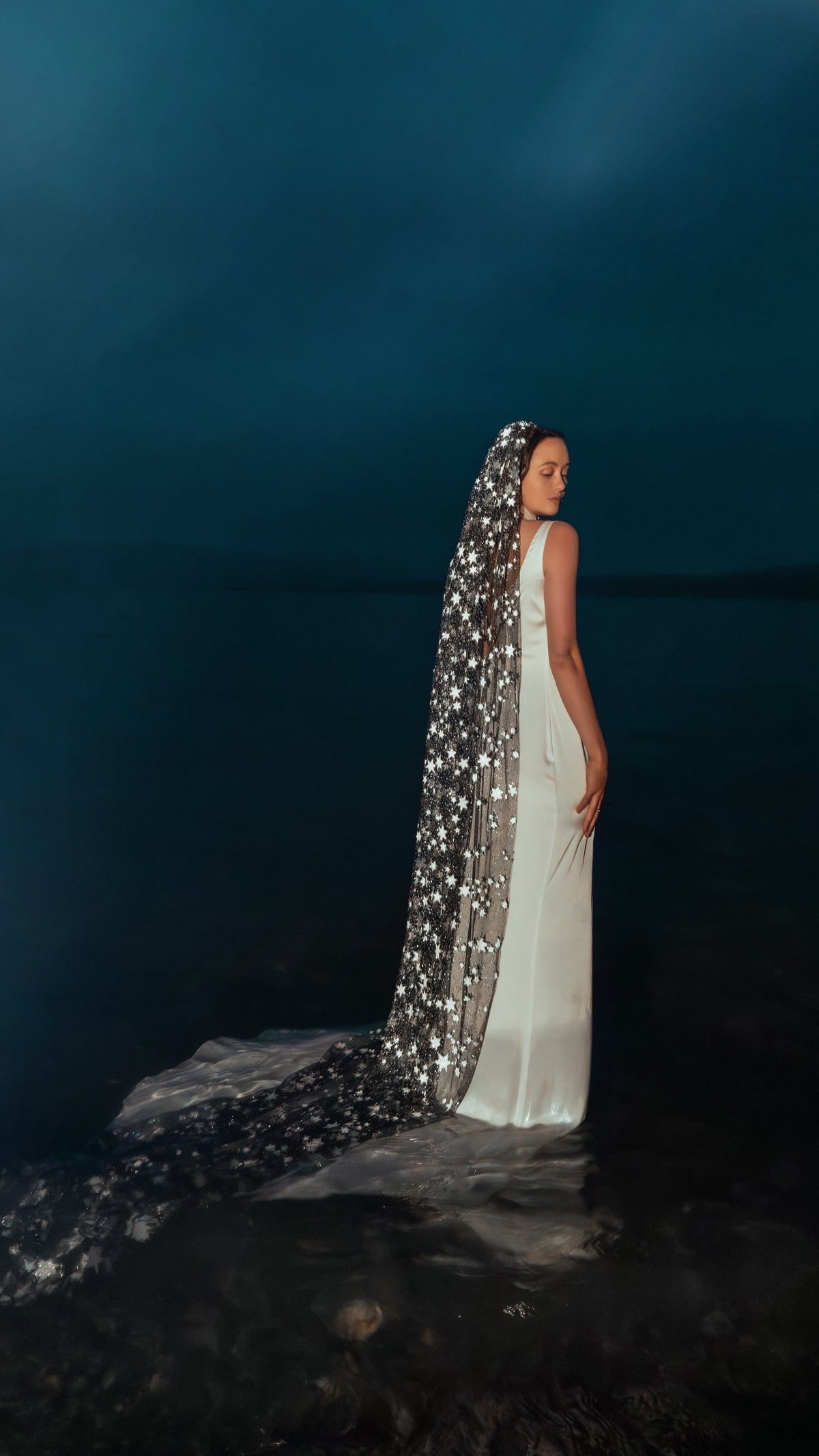 Bride standing in a lake at blue dusk, star wedding veil trailing on the water behind a satin slip dress, Milky Way Veil