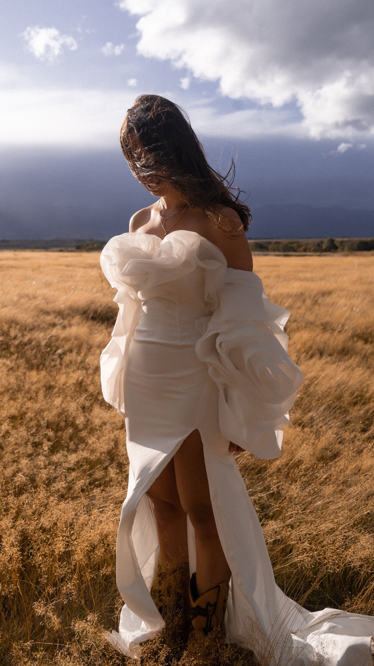Bride with head bowed in a high-slit gown and petal organza detachable sleeves, cowboy boots, open grassland, My Darling Hurricane Sleeves