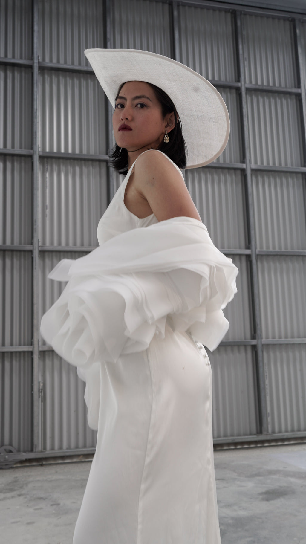 Waist-up three-quarter view of an organza petal bridal jacket worn low on the arms, wide-brim hat, grey warehouse wall, My Darling Hurricane