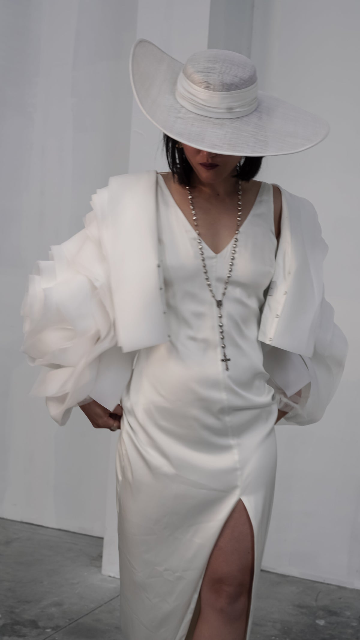 Petal-sleeve bridal jacket over a V-neck slit slip dress and long beaded necklace, hat brim shading the face, My Darling Hurricane