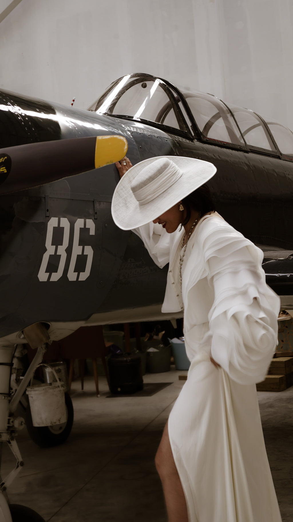 Bridal jacket with petal sleeves over a white slip dress, model in a wide-brim hat beside an old fighter plane in a hangar, My Darling Hurricane